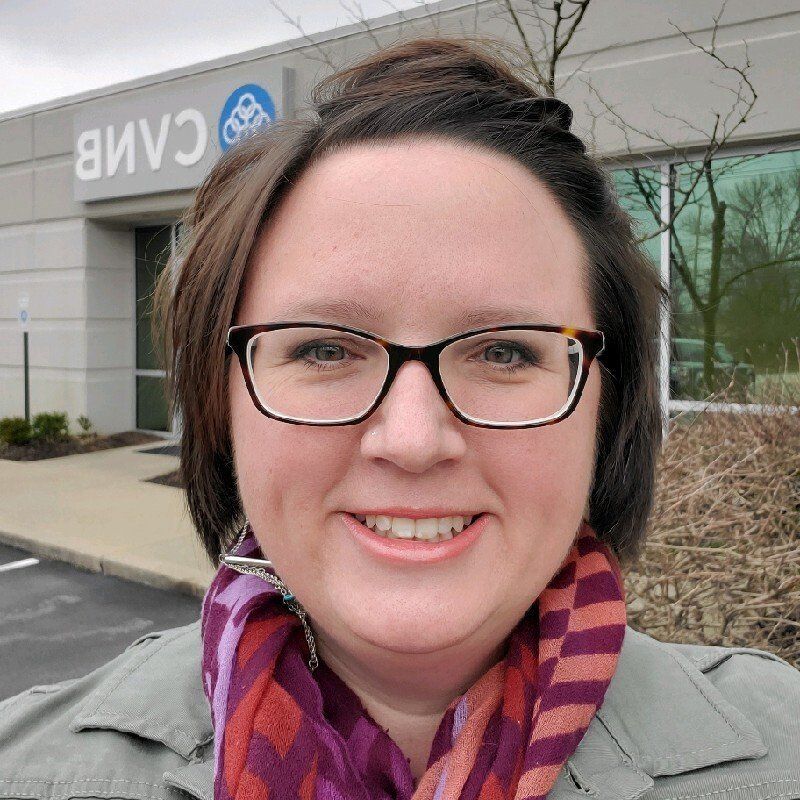 A woman wearing glasses and a scarf is smiling in front of a building that says camb