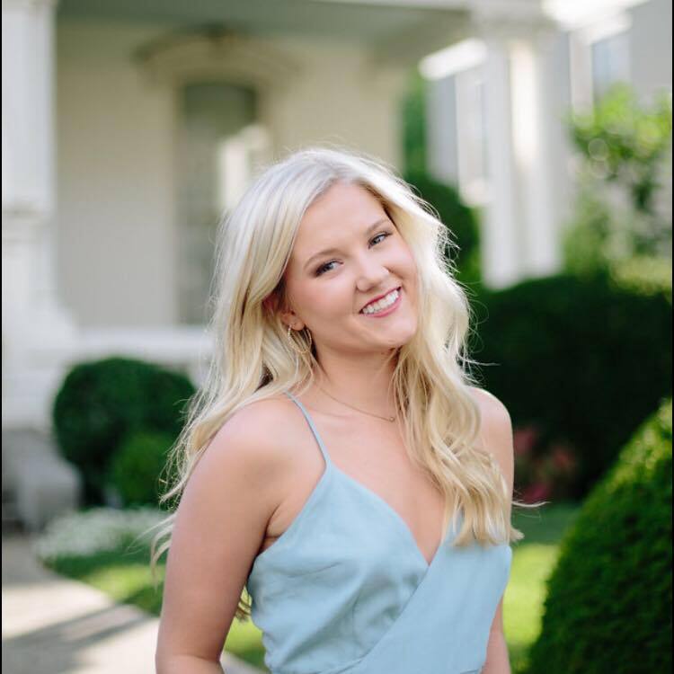A woman in a blue dress is smiling in front of a house.