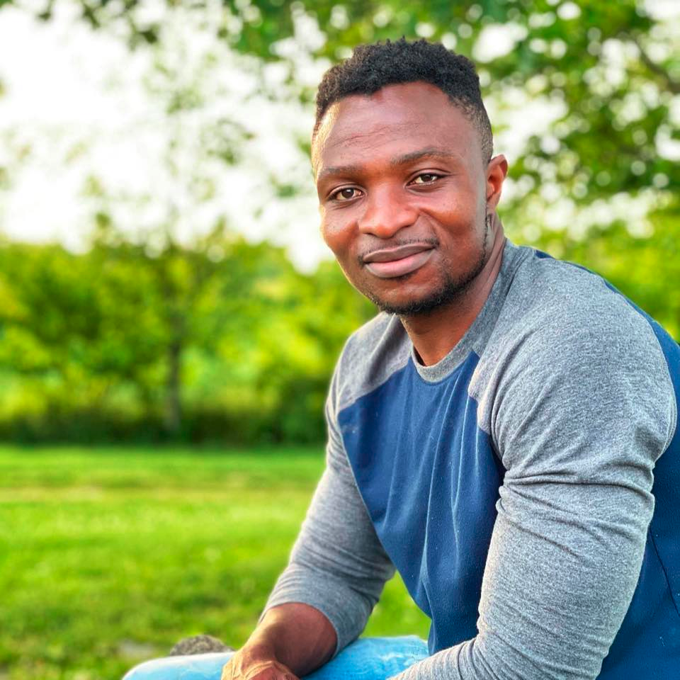 A man in a blue shirt is sitting in the grass and smiling.