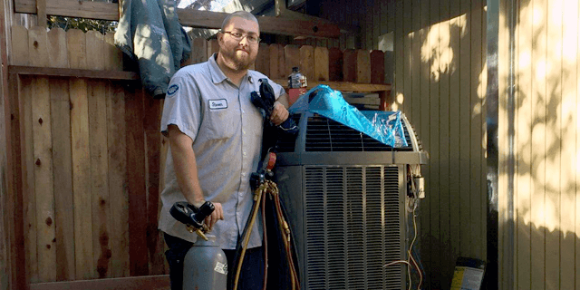 A man is standing in front of an air conditioner holding a hose.