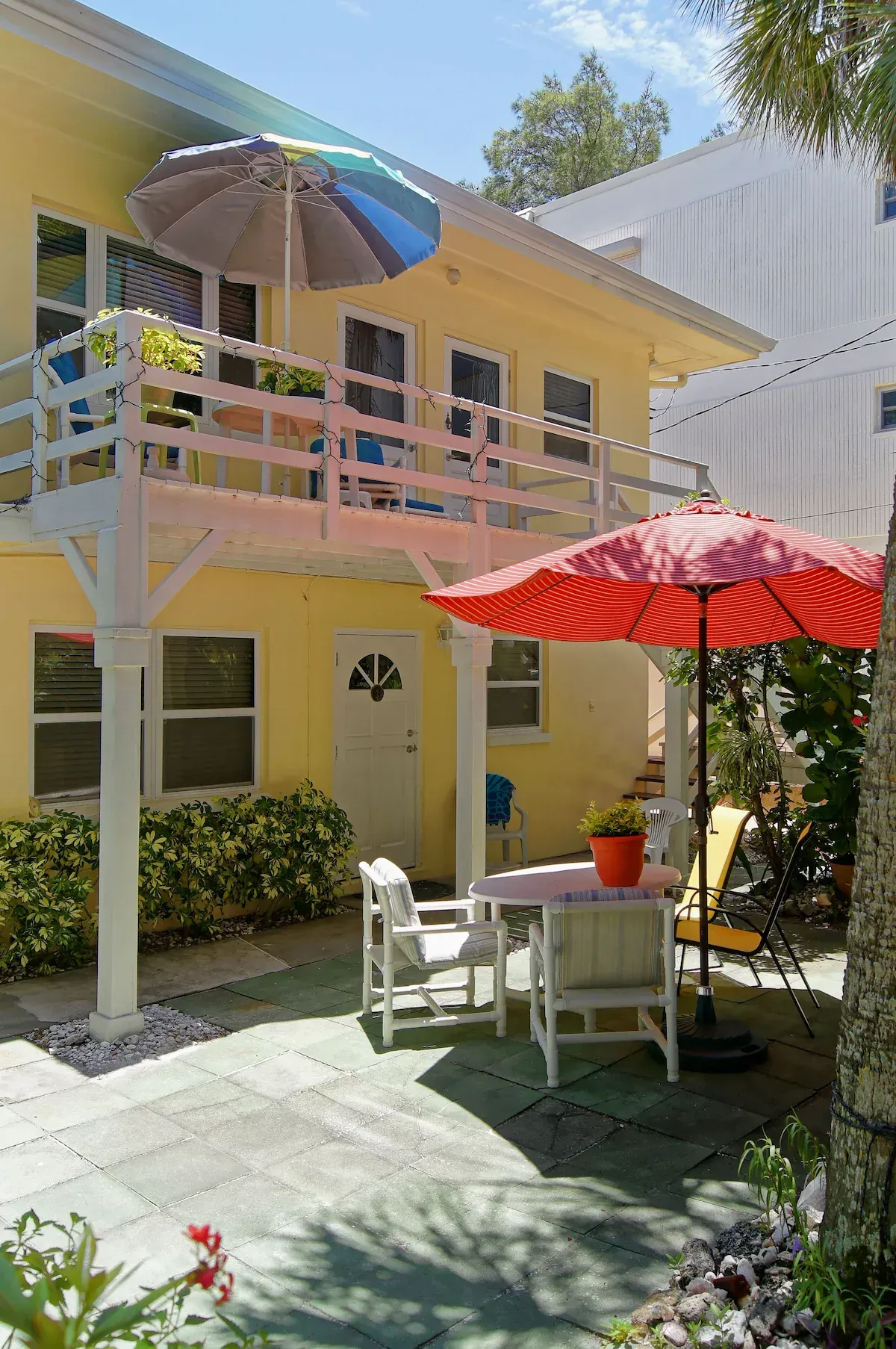 Sunny courtyard with yellow two-story house, patio table, and colorful umbrellas in a small garden courtyard