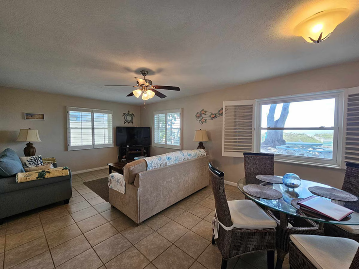 Bright living room with sofa, tiled floor, ceiling fan, TV, and large windows overlooking water