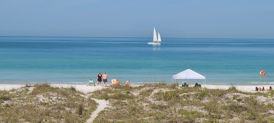 Sailboat on calm blue sea beyond a sandy beach with a few people and a white canopy tent.
