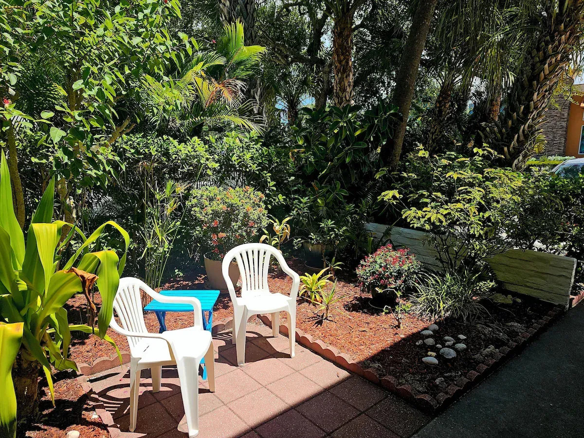 White chairs around a small blue table in a lush garden patio with potted plants and hedges