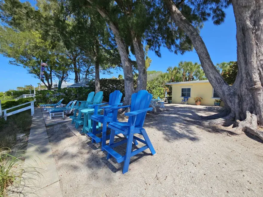 Blue Adirondack chairs under trees beside a sandy path and white fence near a yellow building