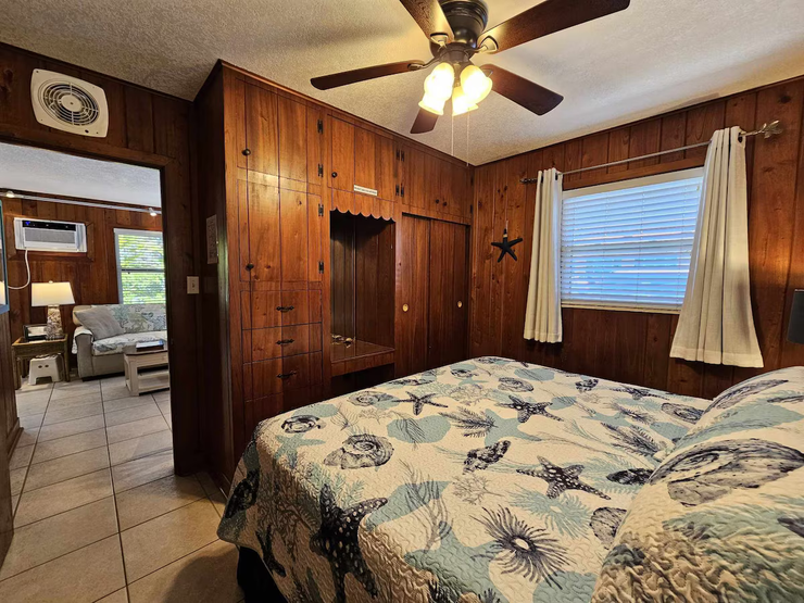 Wood-paneled bedroom with a floral bedspread, ceiling fan, and window with curtains