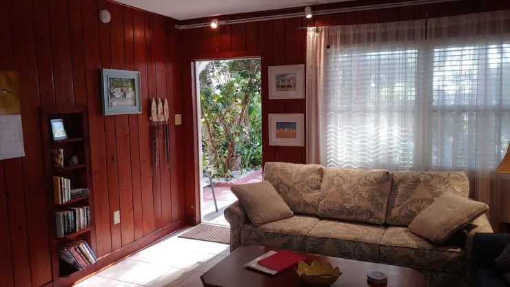 Cozy living room with wood-paneled walls, beige sofa, coffee table, and sunlight through sheer curtains.