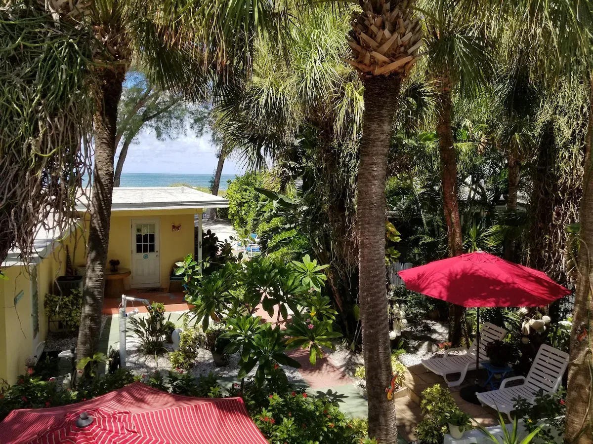 Tropical courtyard with palm trees, yellow building, and red umbrellas near the beach