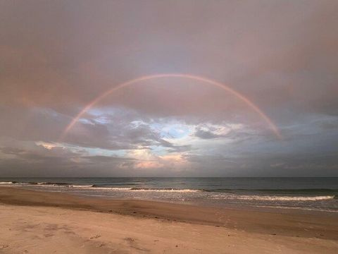 Rainbow arcing over a cloudy beach at sunset, with calm waves and sandy shore