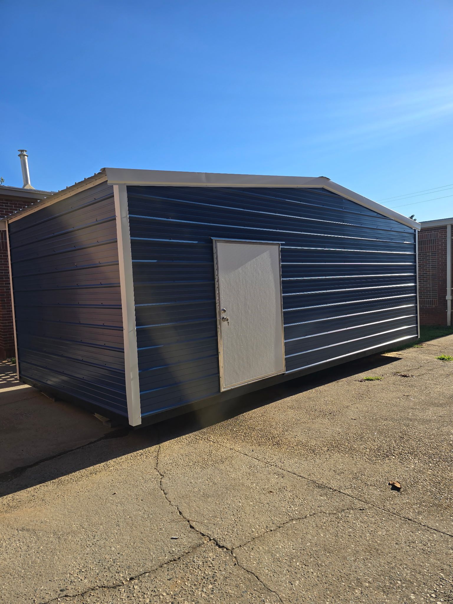 Small dark-blue shed with a white door on a gravel lot under a clear blue sky