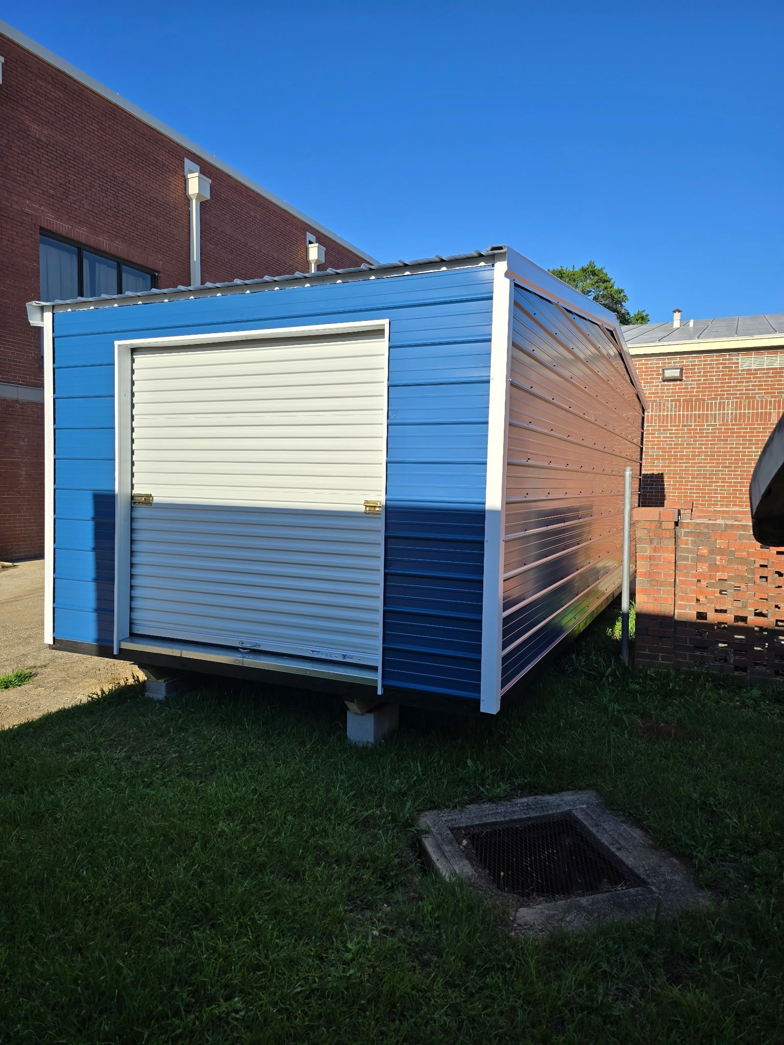 Blue storage shed with a white roll-up door beside a brick building on a grassy lawn.