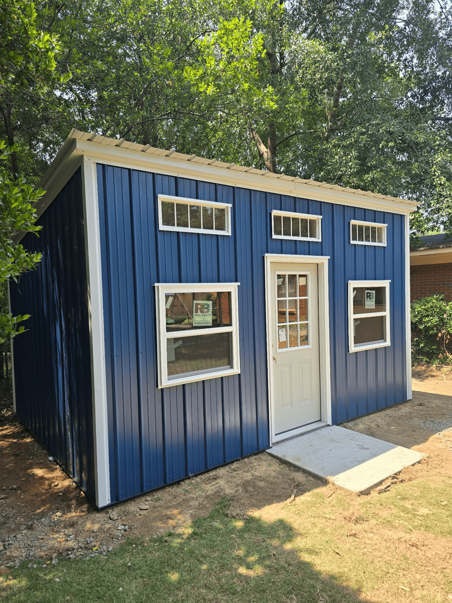 Small blue shed with white trim, windows, and a white door in a sunny wooded yard.
