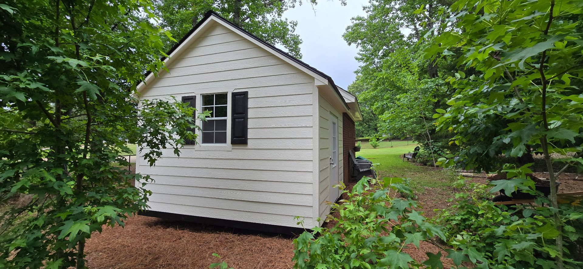 Small white shed with black shutters in a leafy backyard garden path