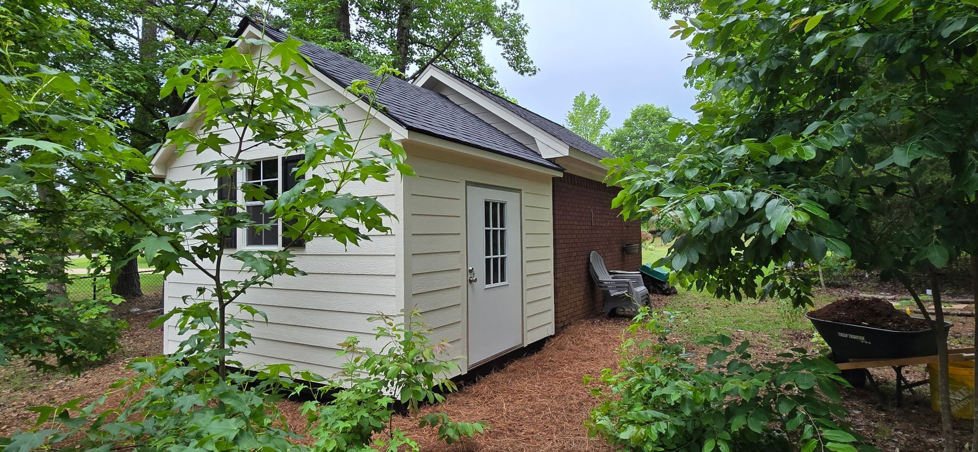 Small white shed with black door in a lush garden, surrounded by trees and mulch path