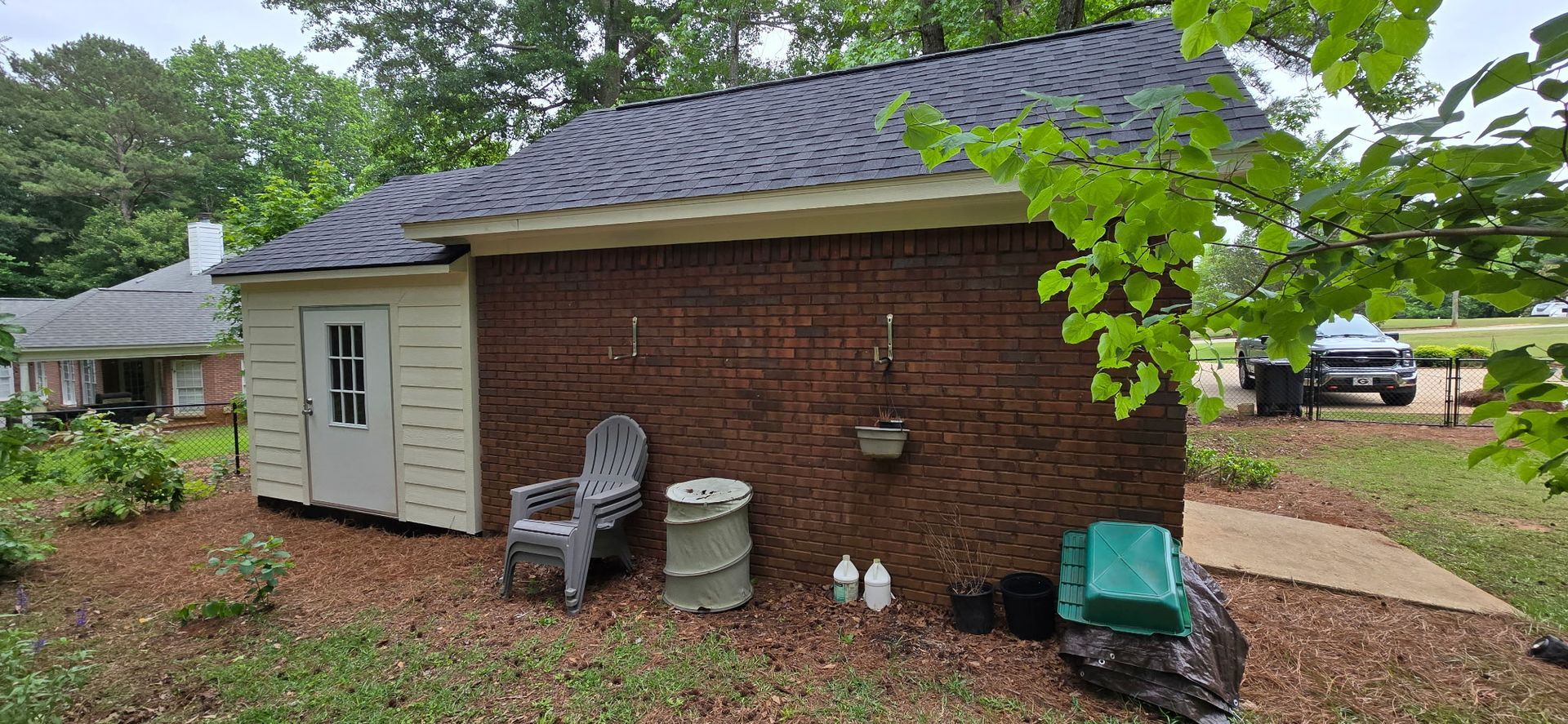 Brick house exterior with white side door, chimney, patio chairs, and yard equipment under trees