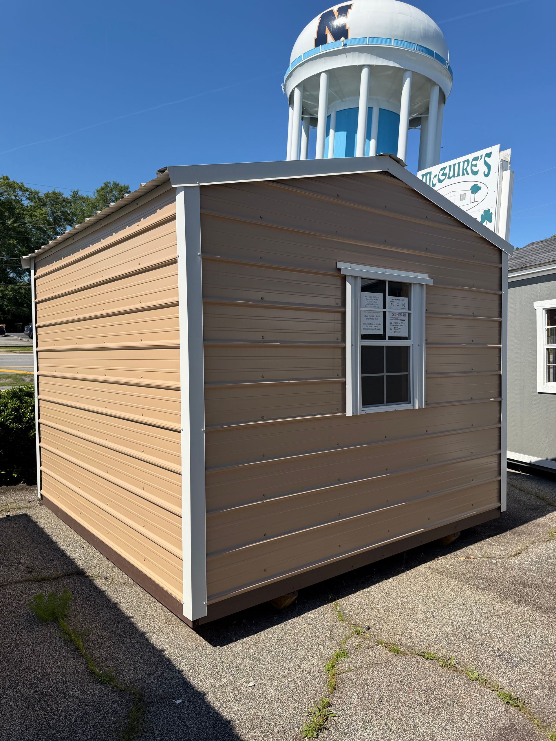 Small tan modular building with white trim and a window, seen outdoors on pavement under a water tower.