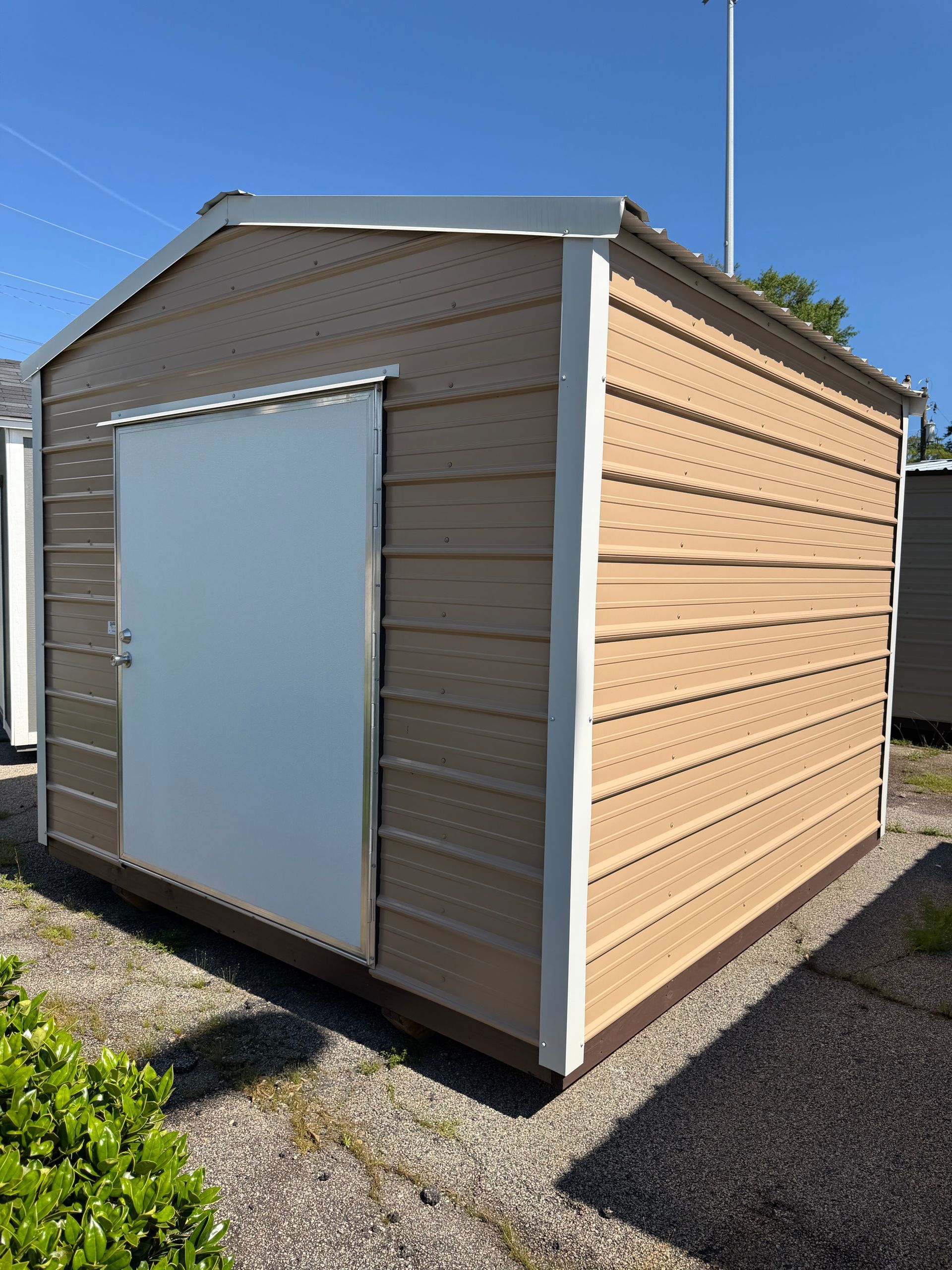 Small tan shed with white trim and a white door, standing on gravel under a clear blue sky