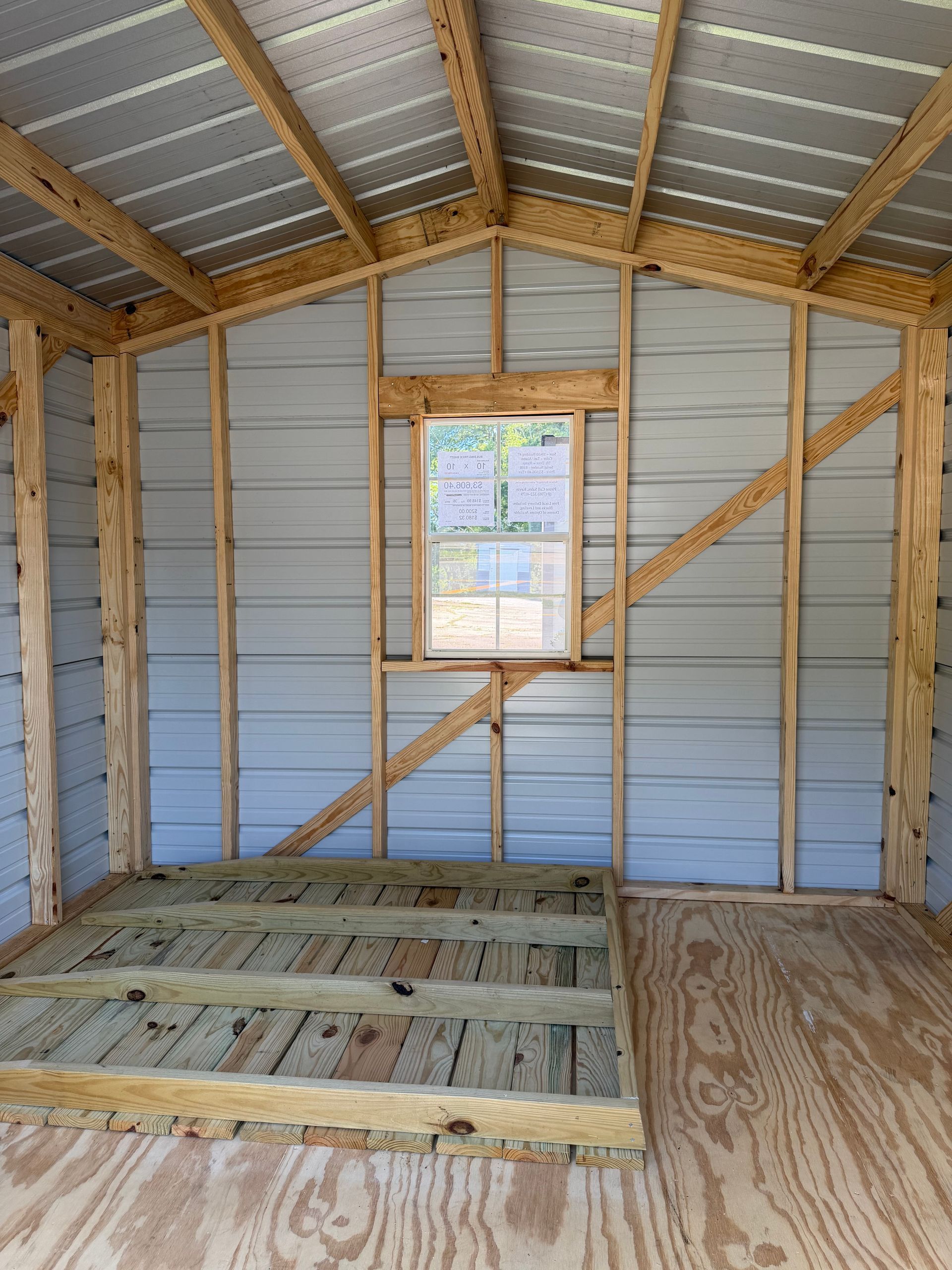 Unfinished wooden shed interior with insulated walls, a small window, and a plywood floor platform.