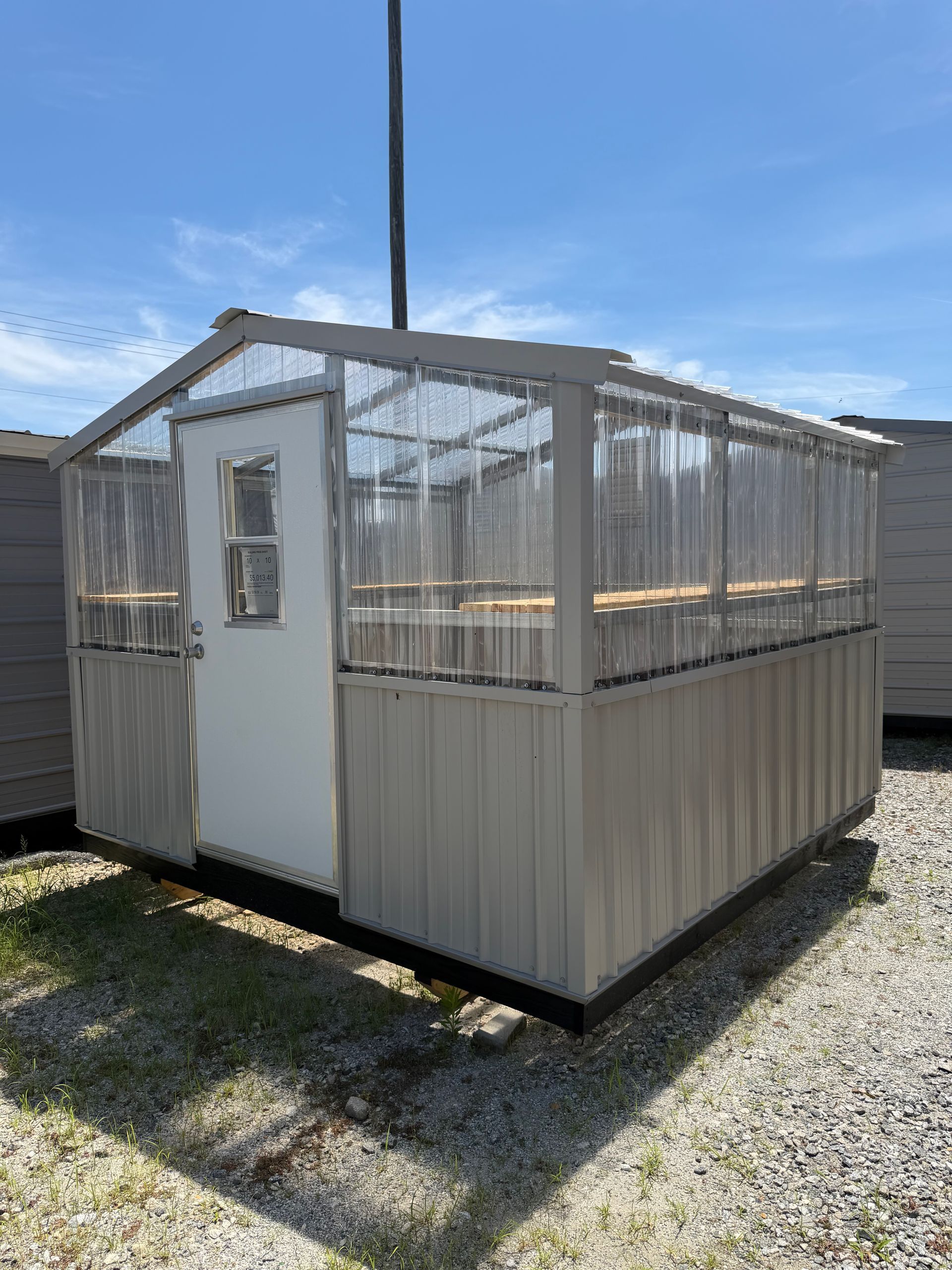 Small white and gray shed with a door and windows, standing on gravel under a blue sky