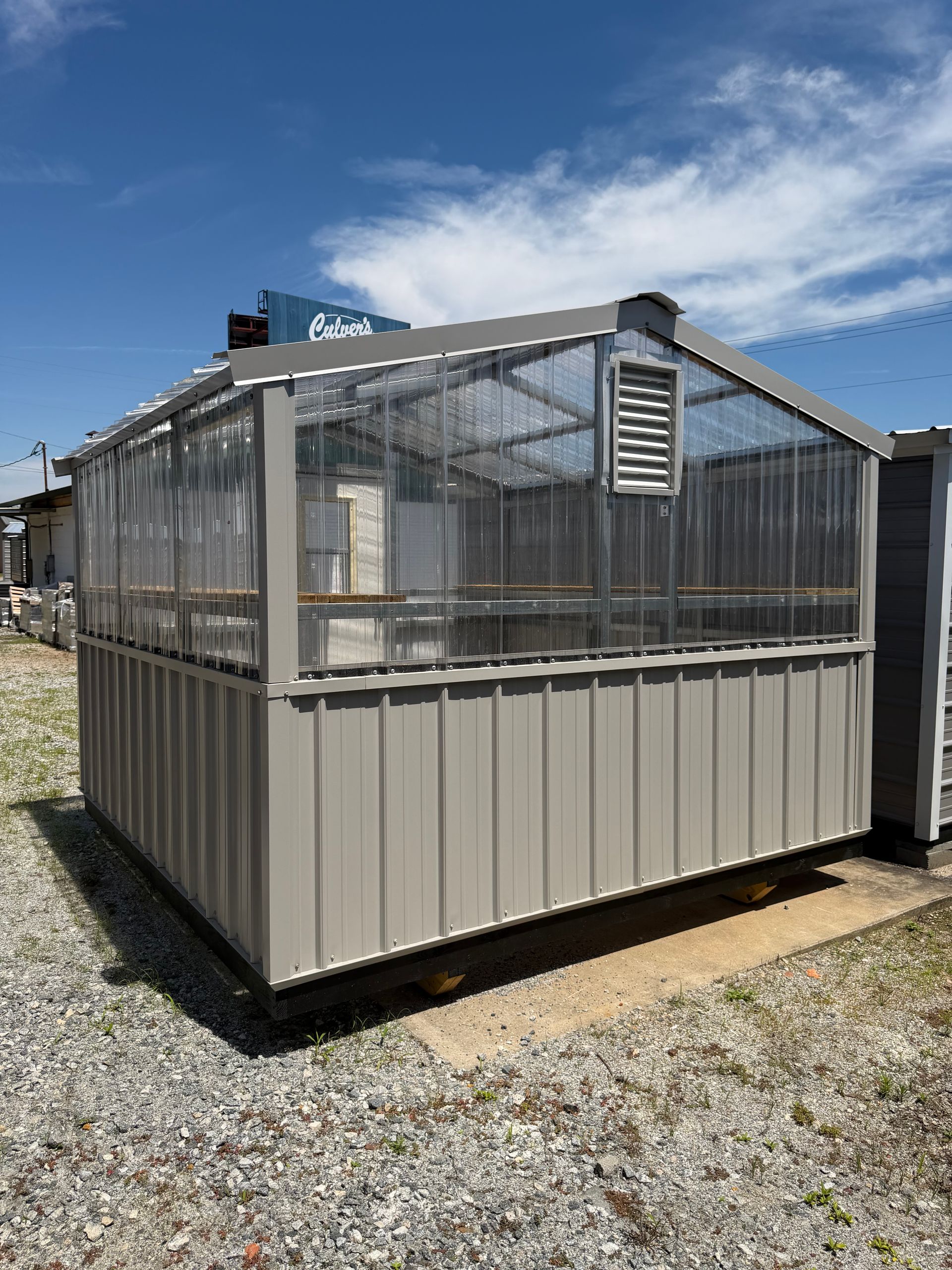 Small beige-and-clear greenhouse on a gravel lot under a blue sky