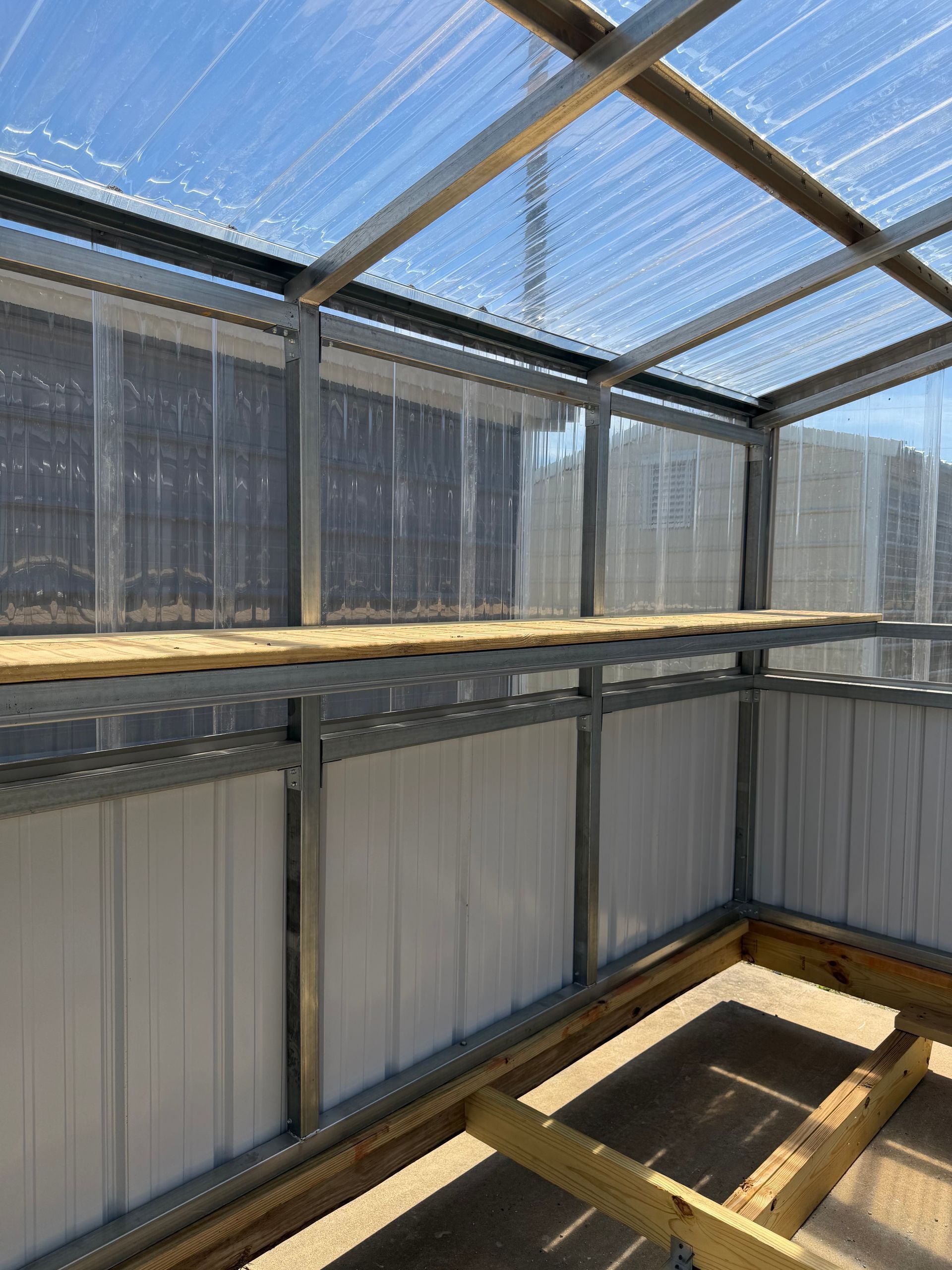 Interior of a greenhouse with metal framing, translucent walls, and a wooden raised bed under a clear roof.