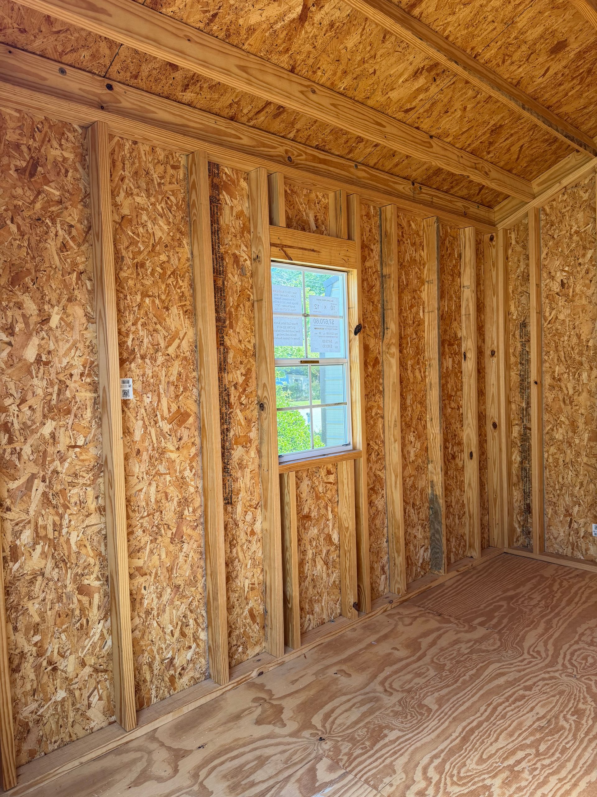 Unfinished wooden shed interior with exposed studs, plywood floor, and a small window showing greenery outside