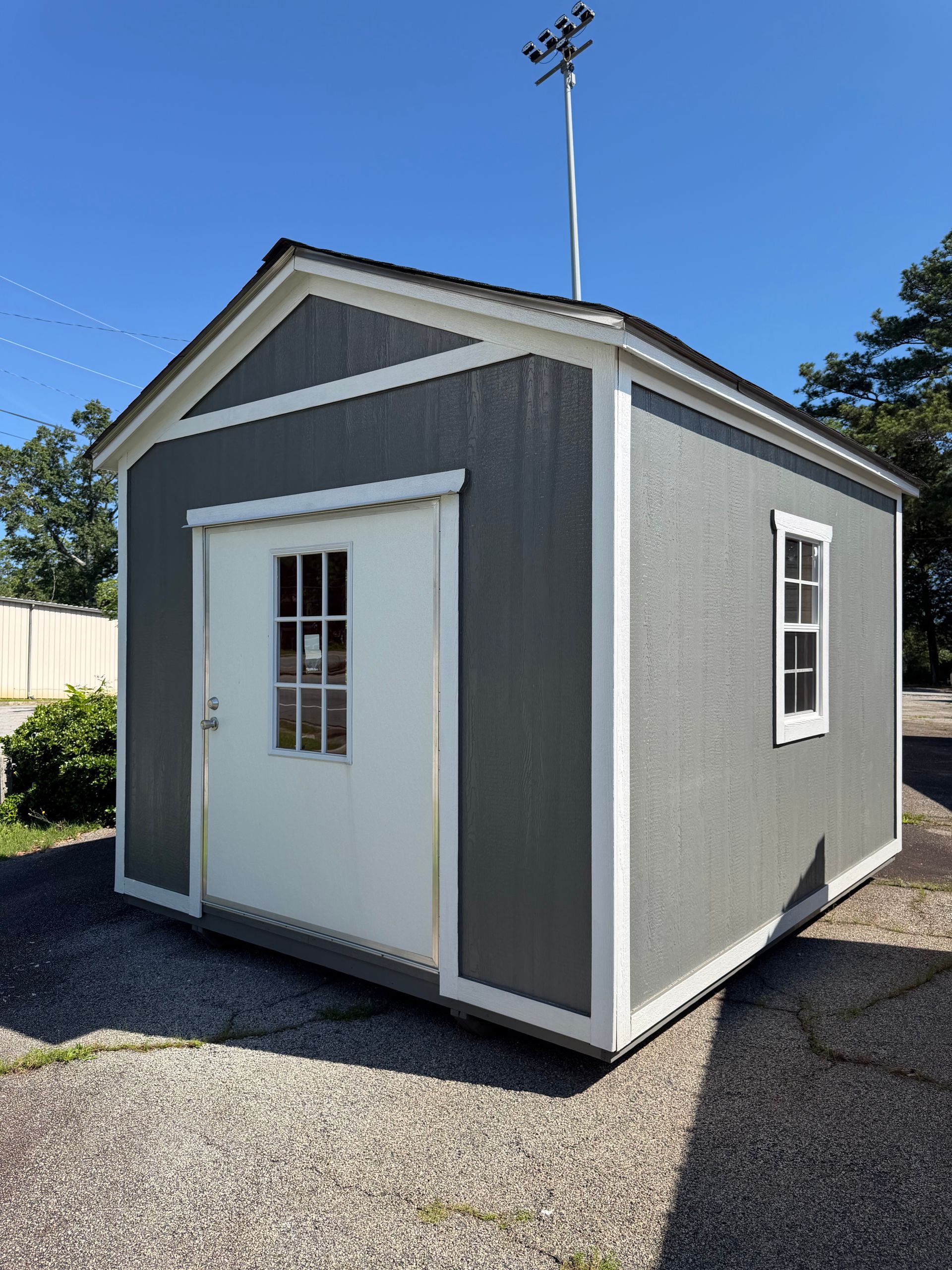 Small gray shed with white trim and a door, sitting on a gravel lot under a blue sky.