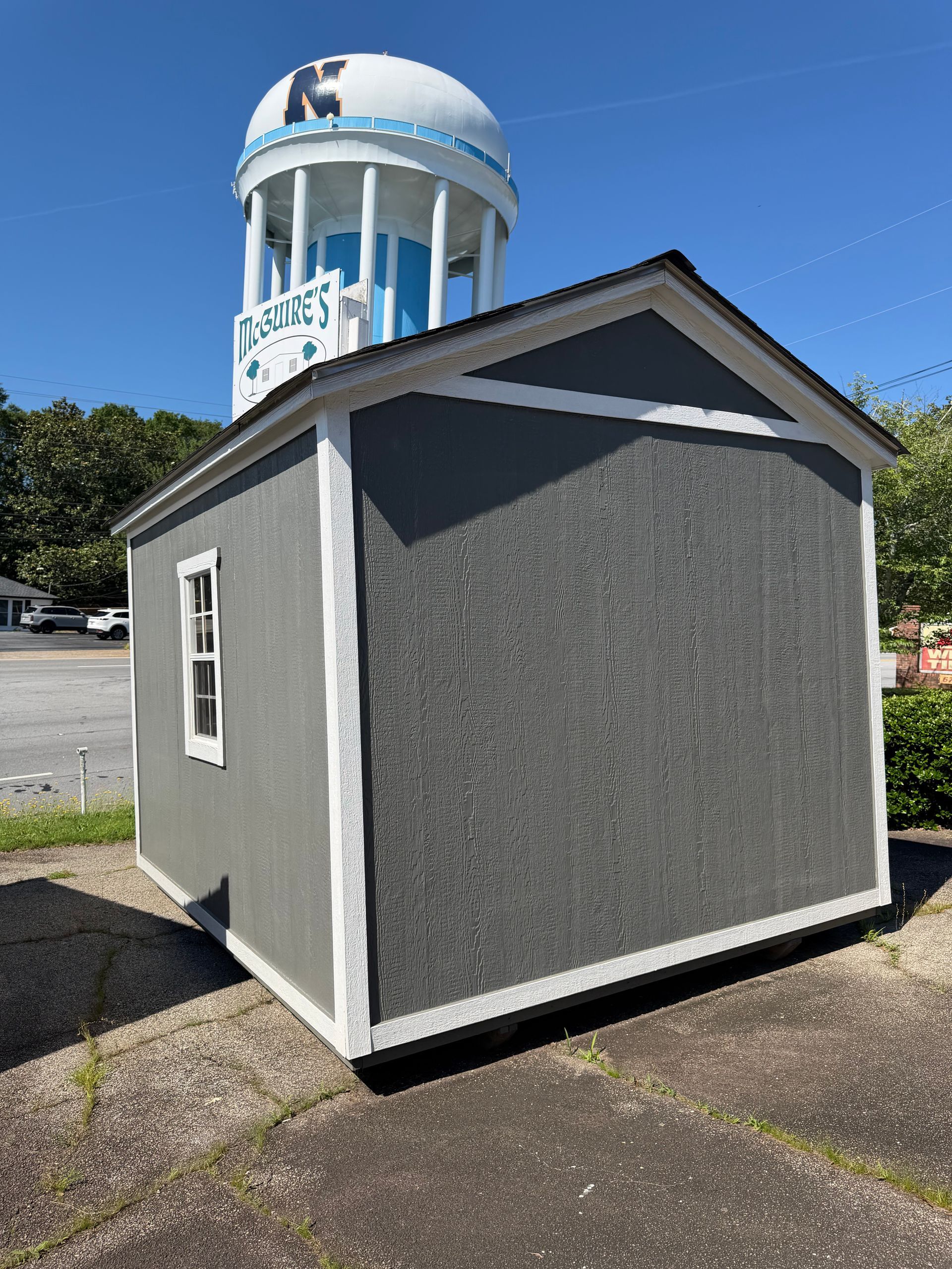Small gray utility shed with white trim in a parking lot, with a blue-and-white water tower behind it.