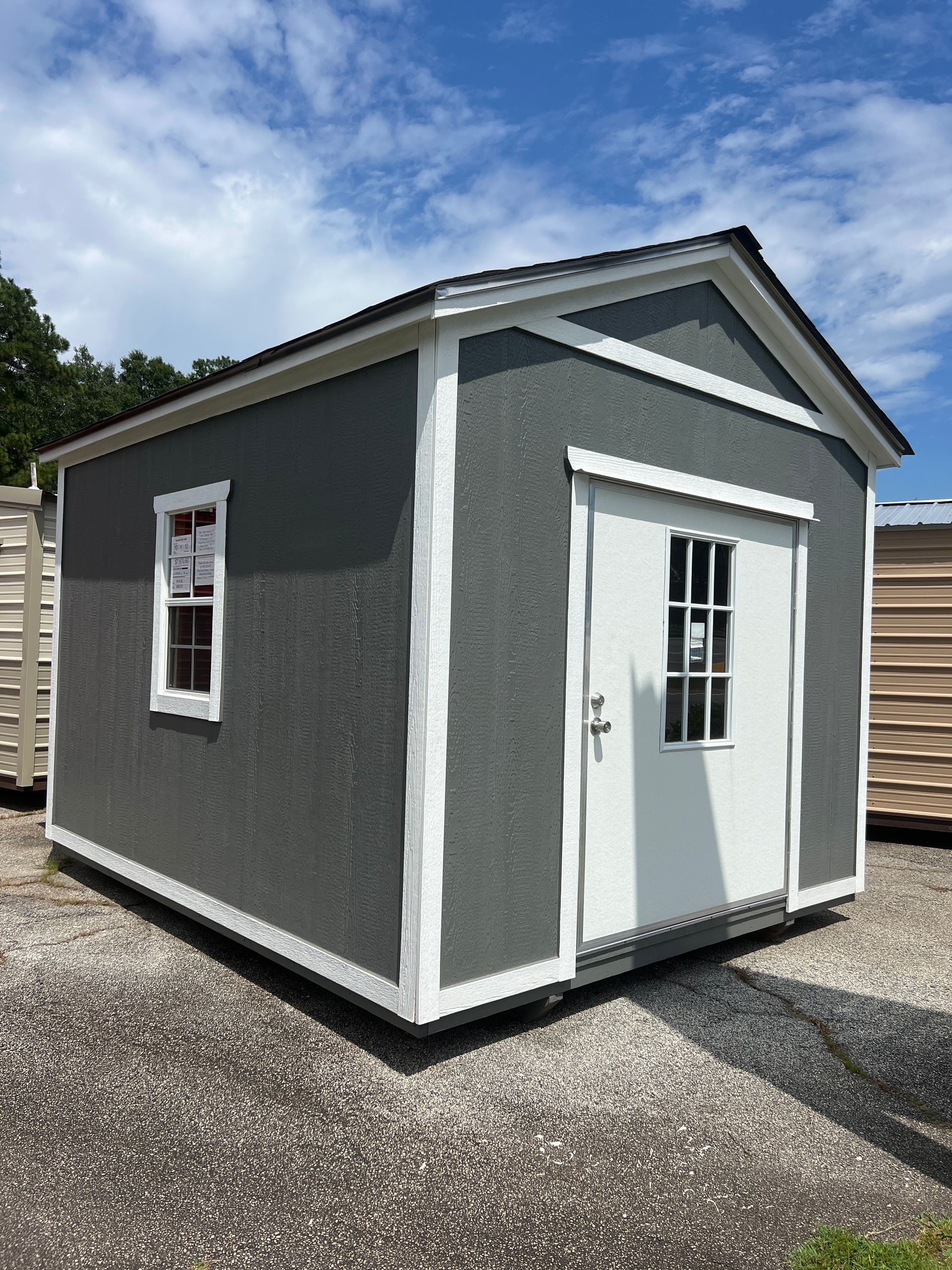 Small gray shed with white trim and a white door on a gravel lot under a blue sky