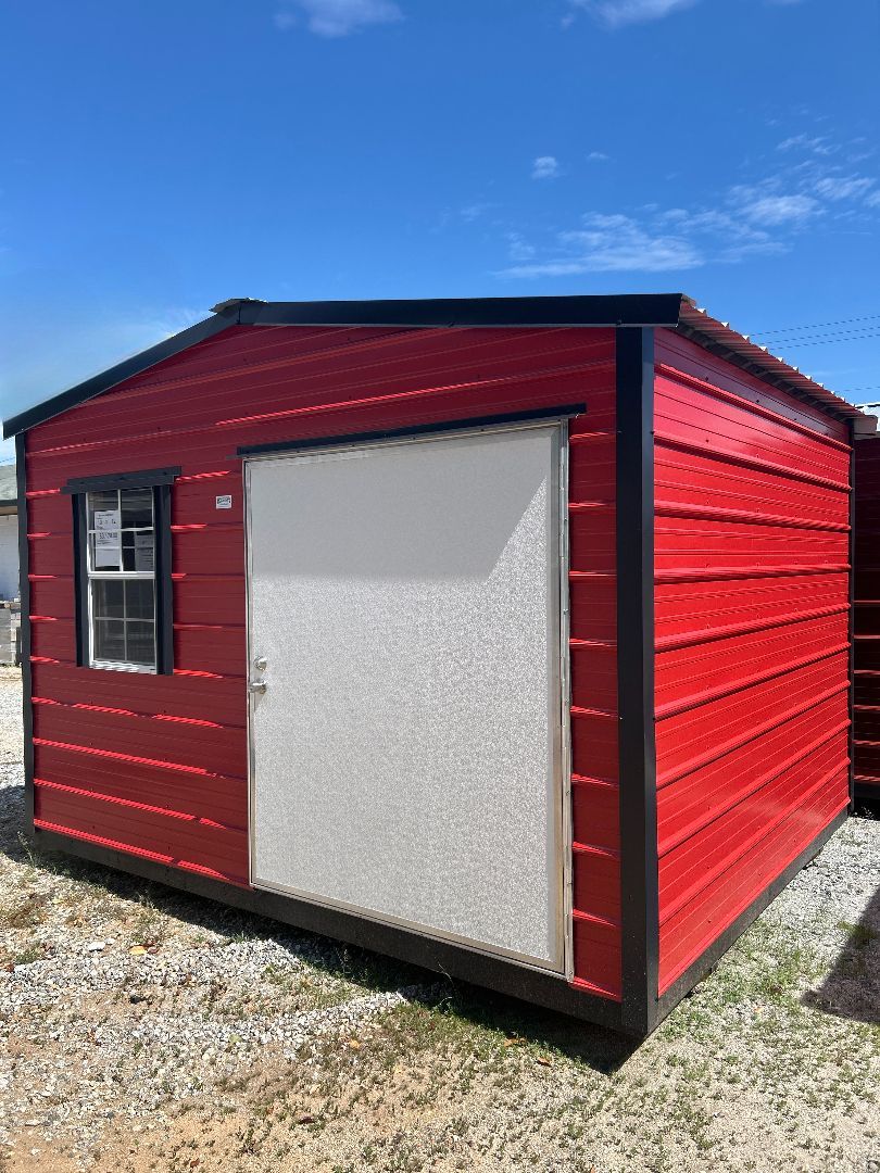 Red shed with a white door and small window, black trim, sitting outside on gravel under a blue sky.