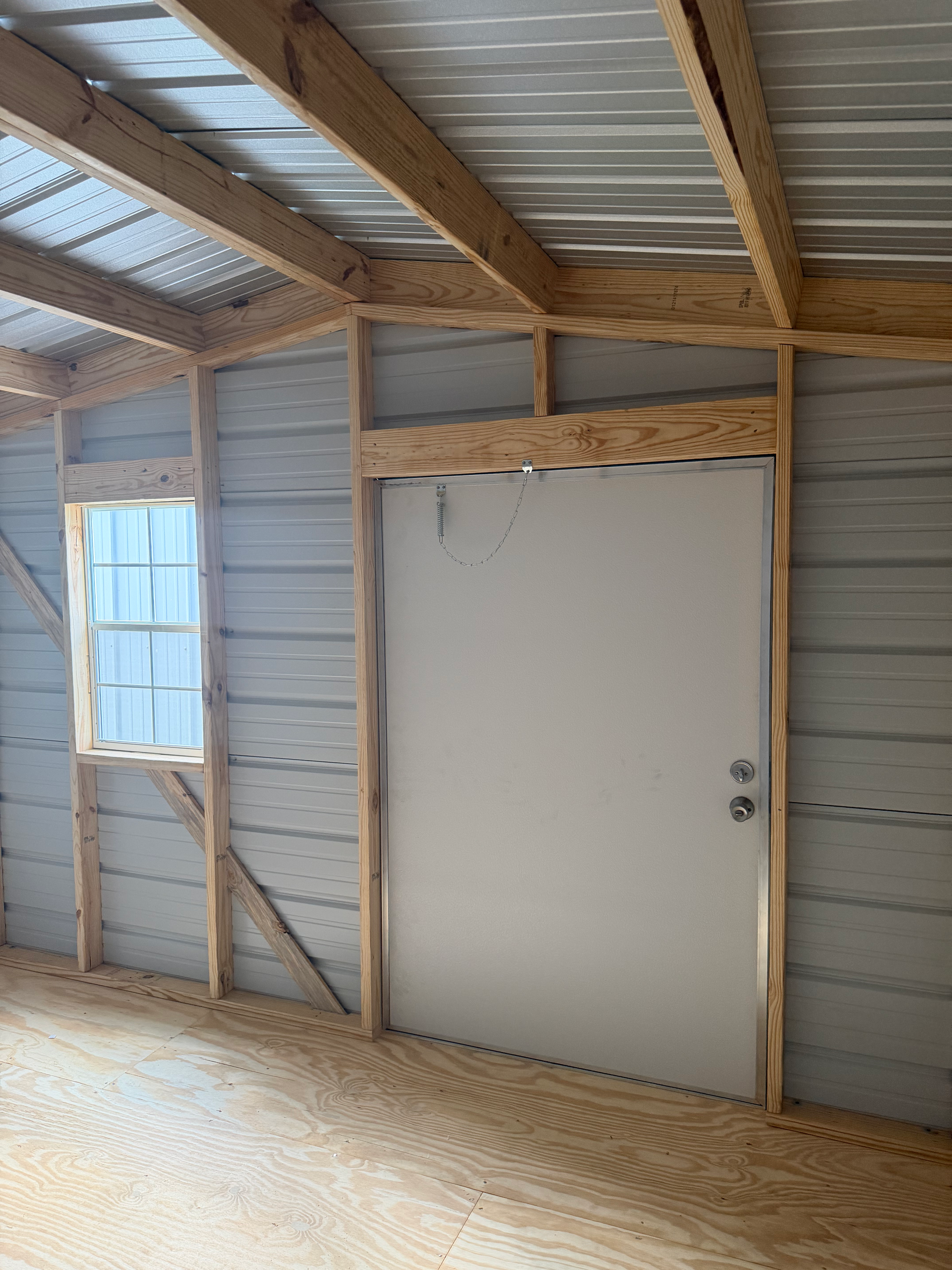 Small unfinished room with wood framing, metal roof, a white door, and a window.