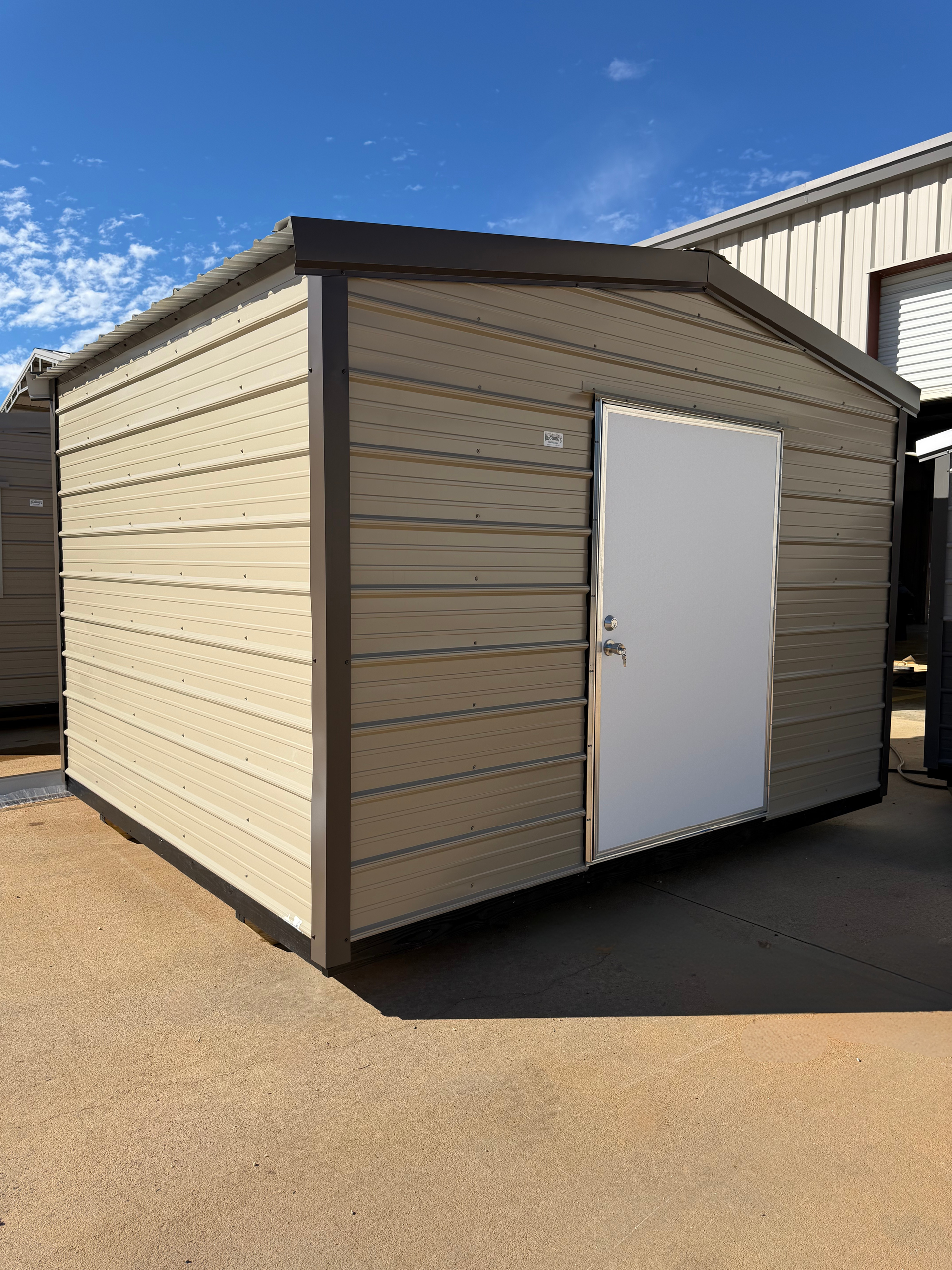 Small beige storage shed with a white door on a sunny paved lot