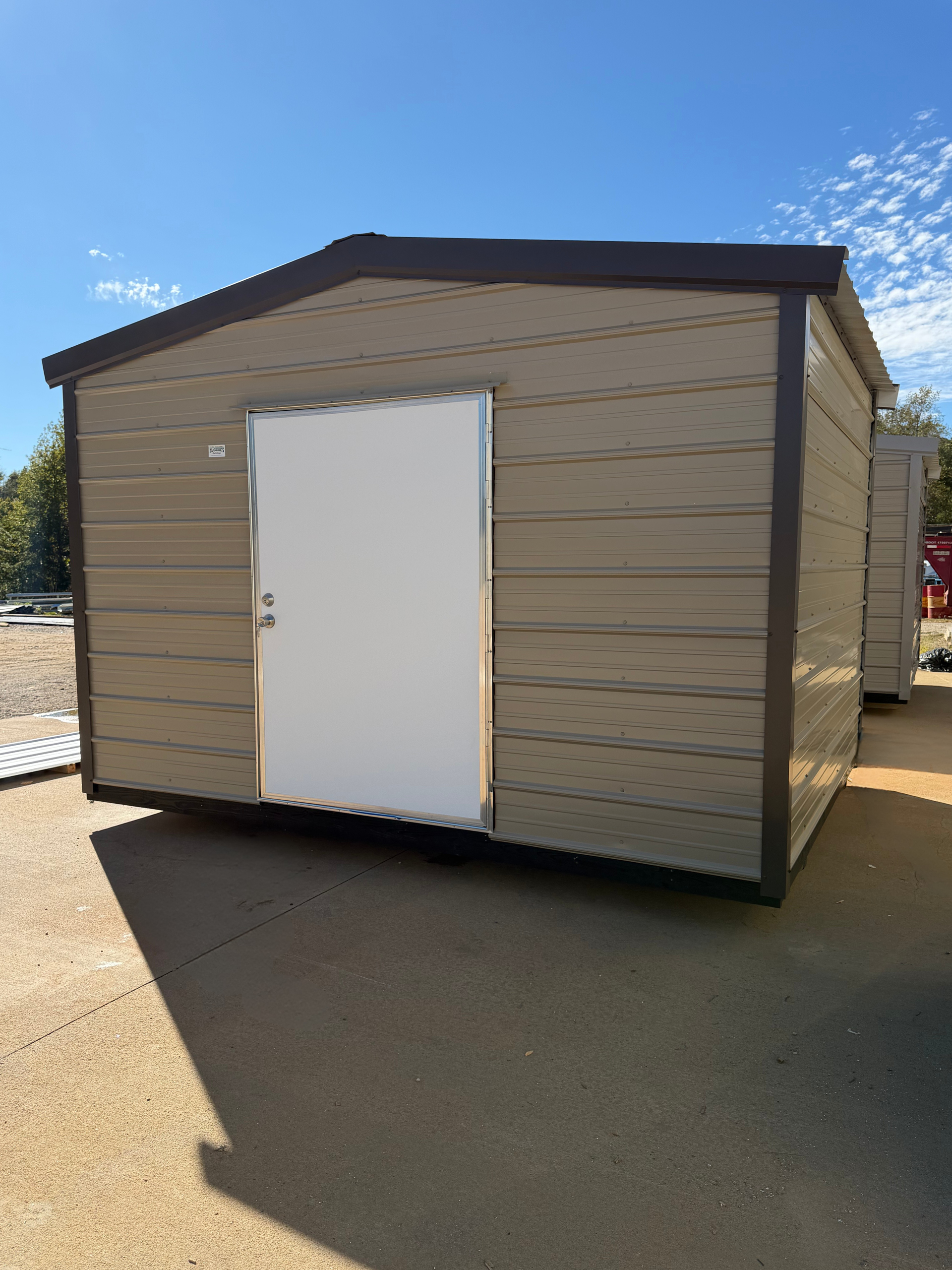 Small tan storage shed with a white door on a sunny paved lot