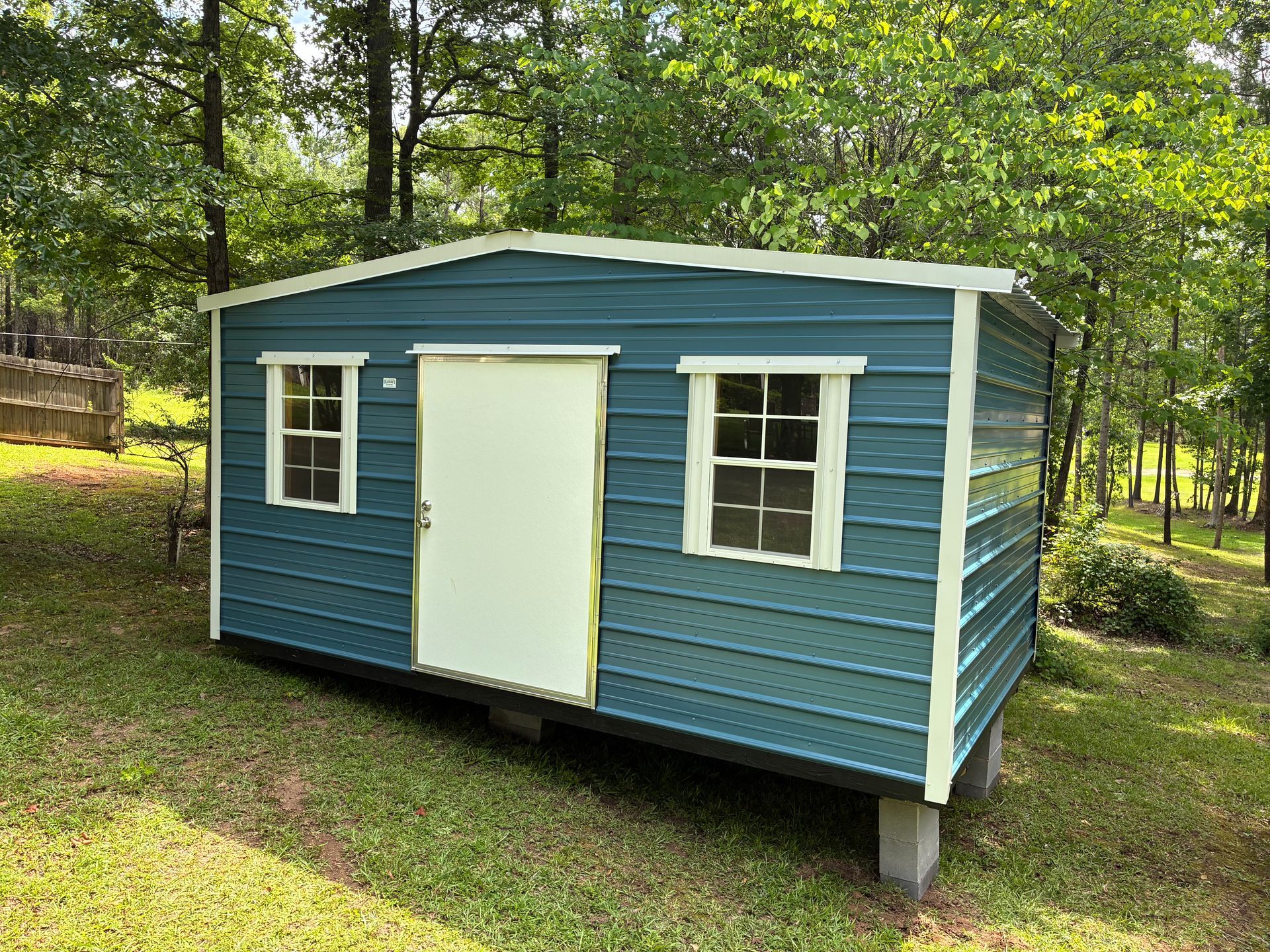 Blue small shed with white door and windows on concrete blocks in a wooded yard
