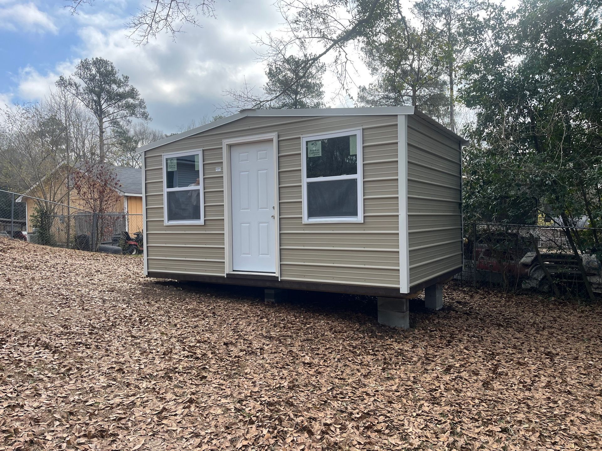 Tan metal shed with white door and windows, set on concrete blocks, outdoors with leaf-covered ground.