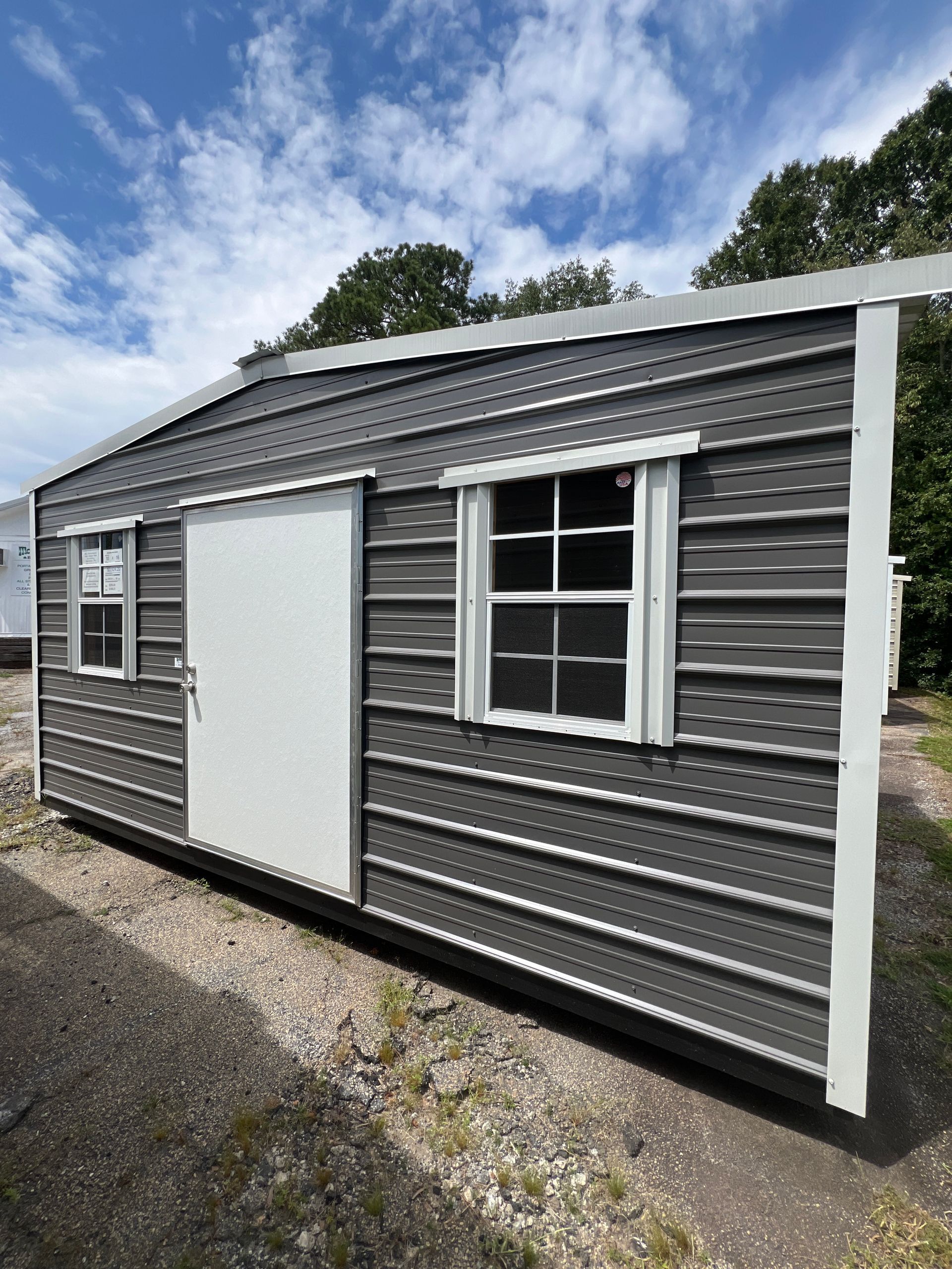 Gray metal shed with white door and window, shown outdoors on a gravel lot under a cloudy sky