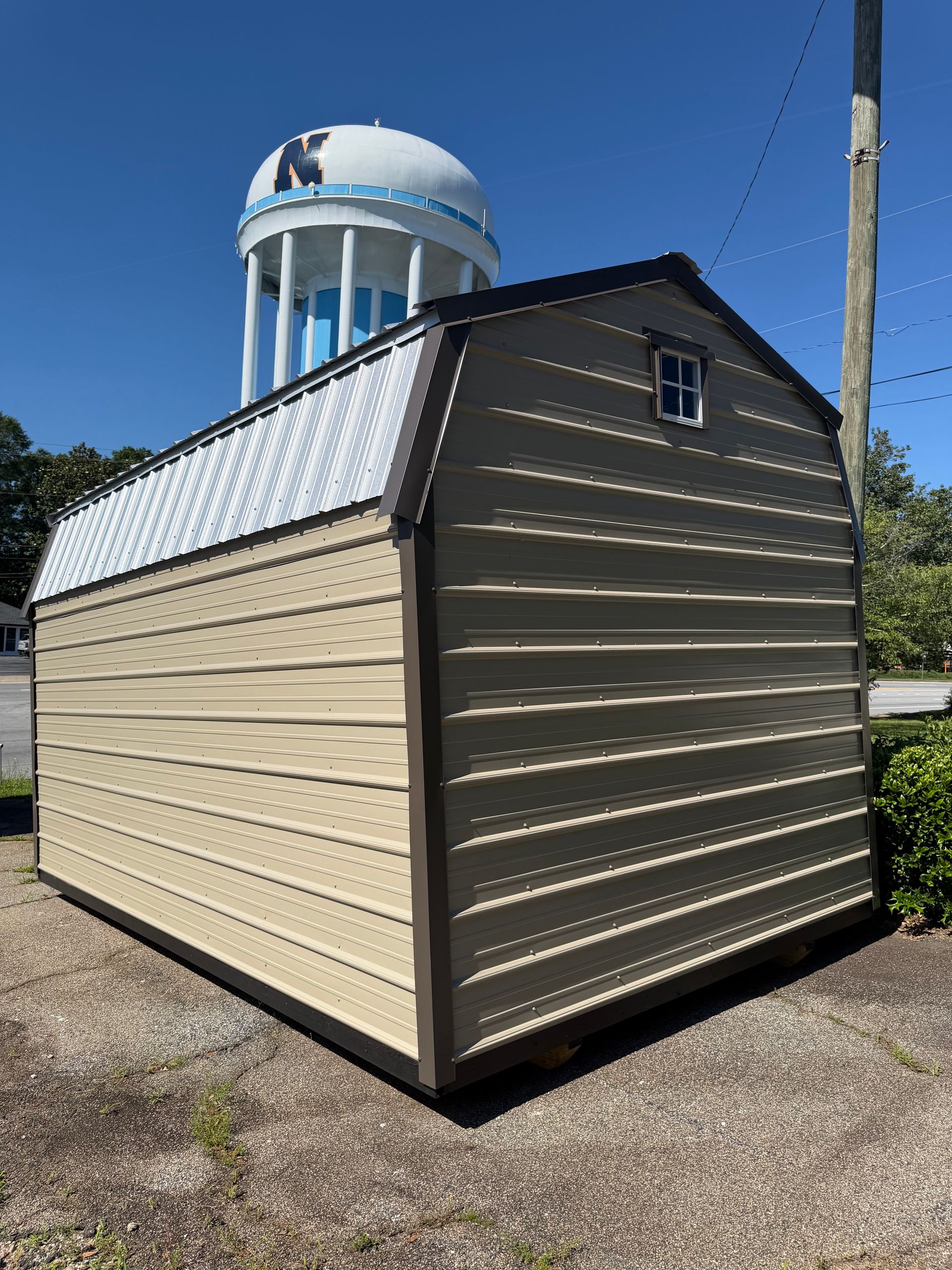 Beige shed with a white metal roof, outdoors on gravel, with a water tower in the background
