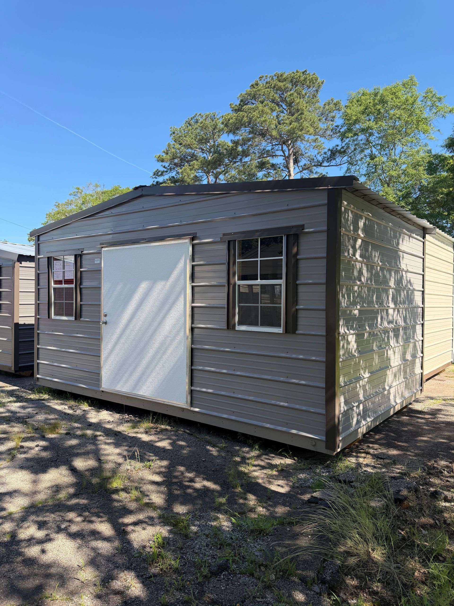 Small gray shed with white door and windows, sitting outdoors on a sunny gravel lot under blue sky.