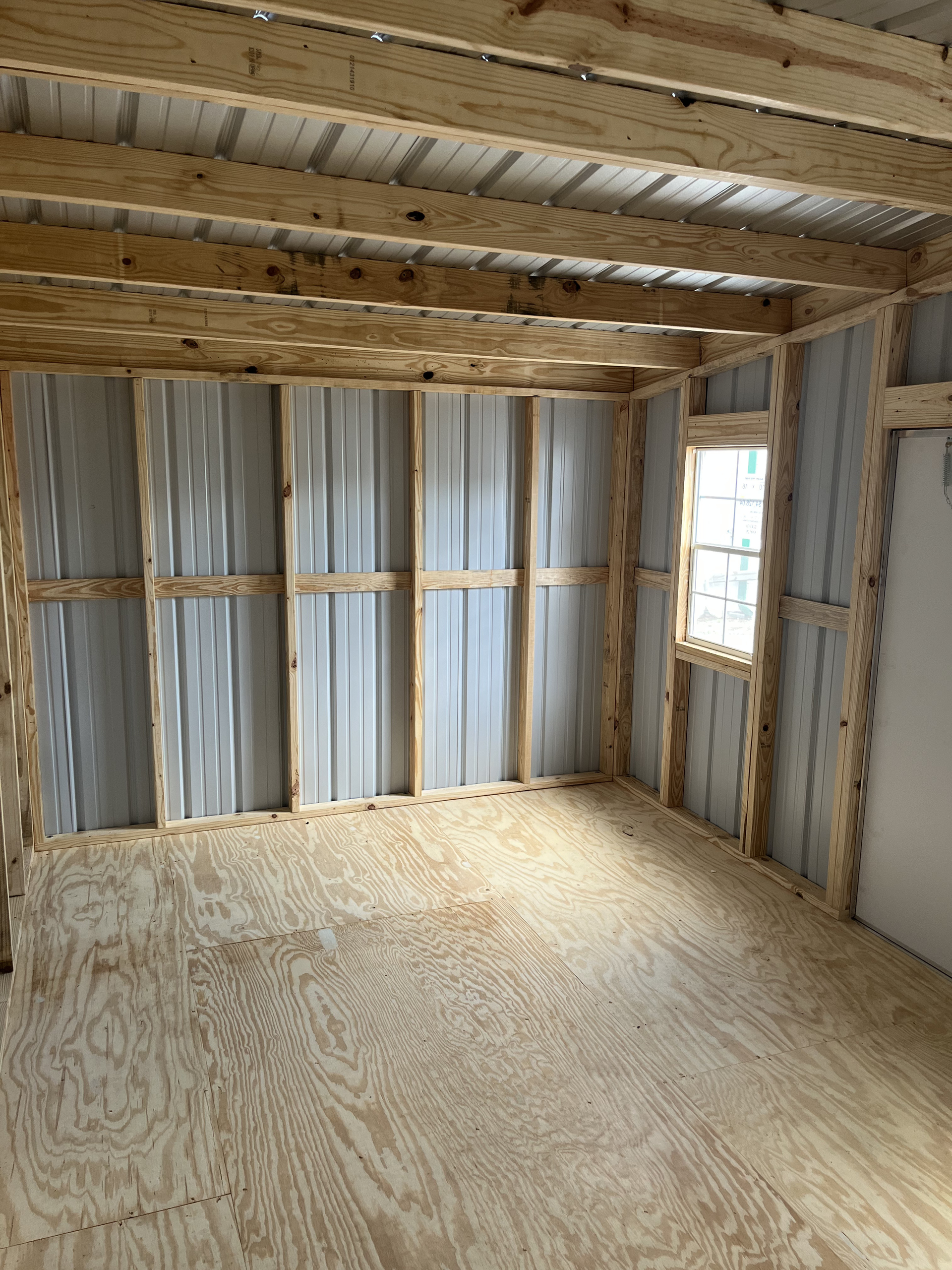 Empty unfinished room with exposed wood framing, plywood floor, and a small window.