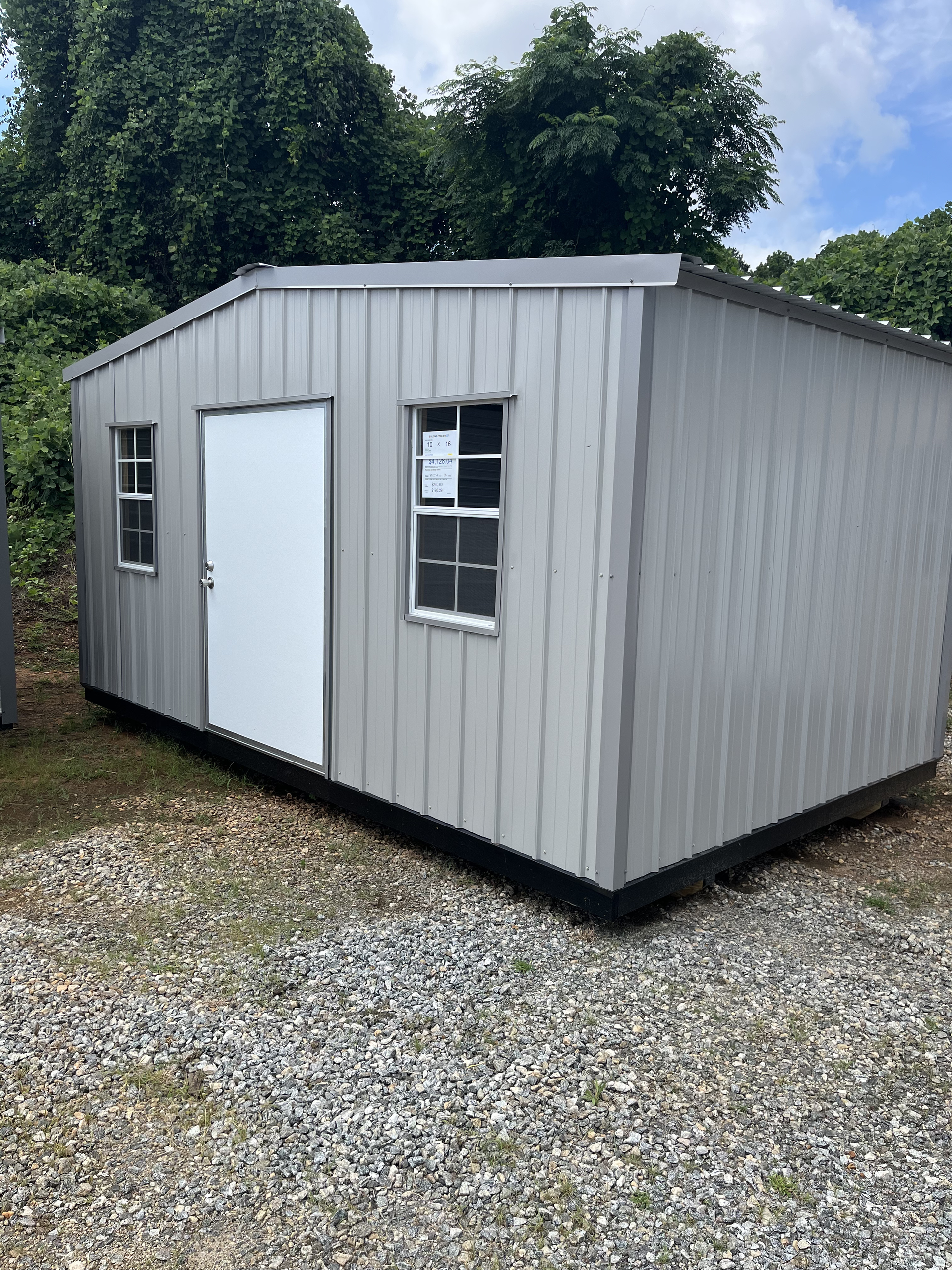 Gray portable shed with a white door and window on a gravel lot, backed by trees under a cloudy sky