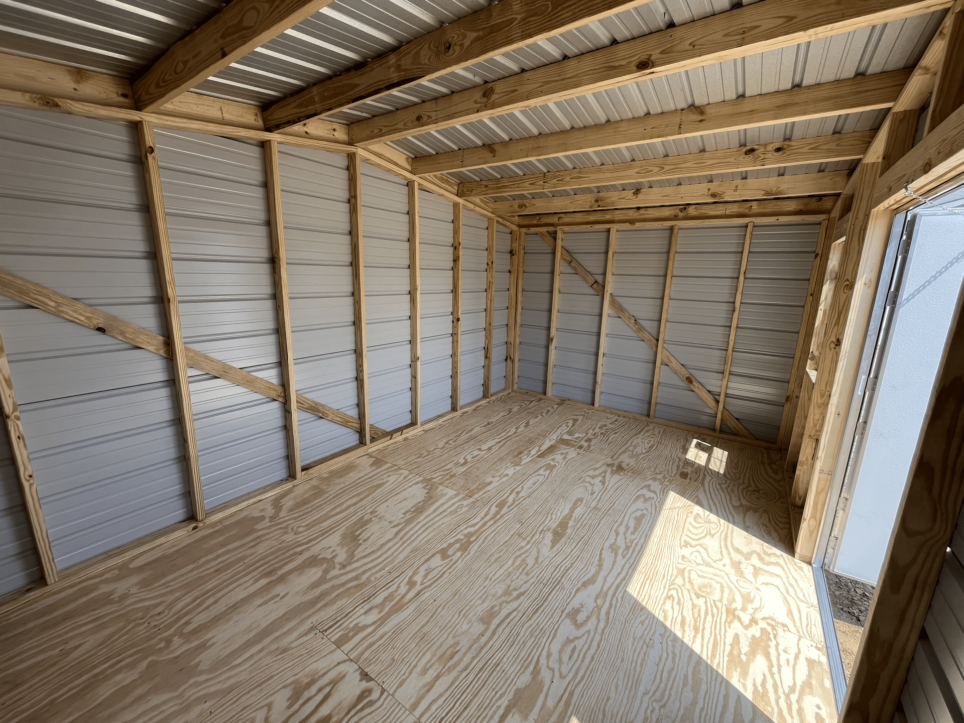 Empty unfinished wooden shed interior with exposed framing, plywood floor, and sunlight through a door opening