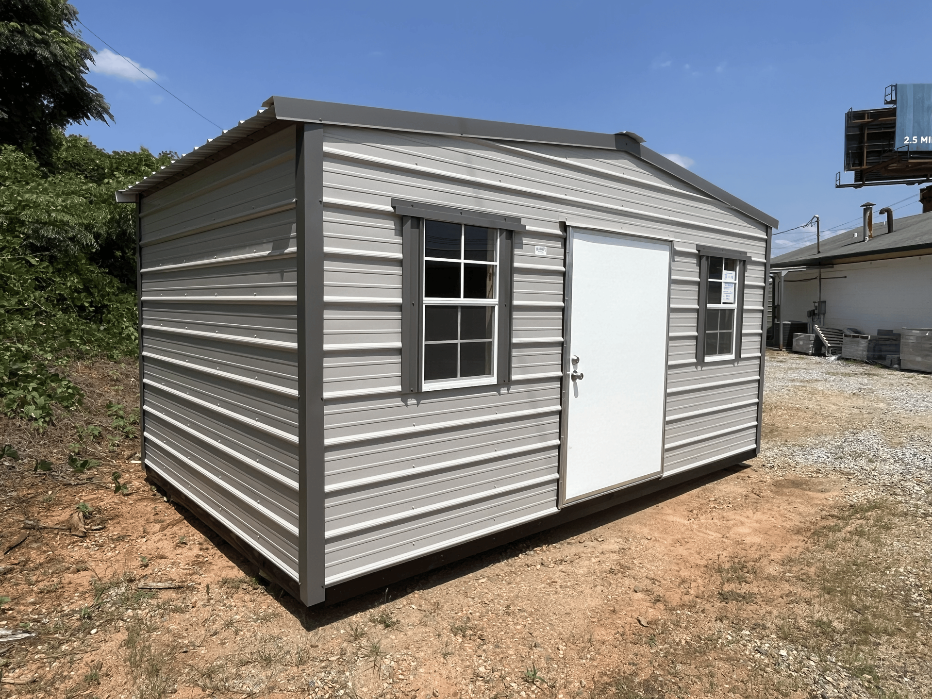 Small gray metal shed with white door and windows on a gravel lot under a blue sky