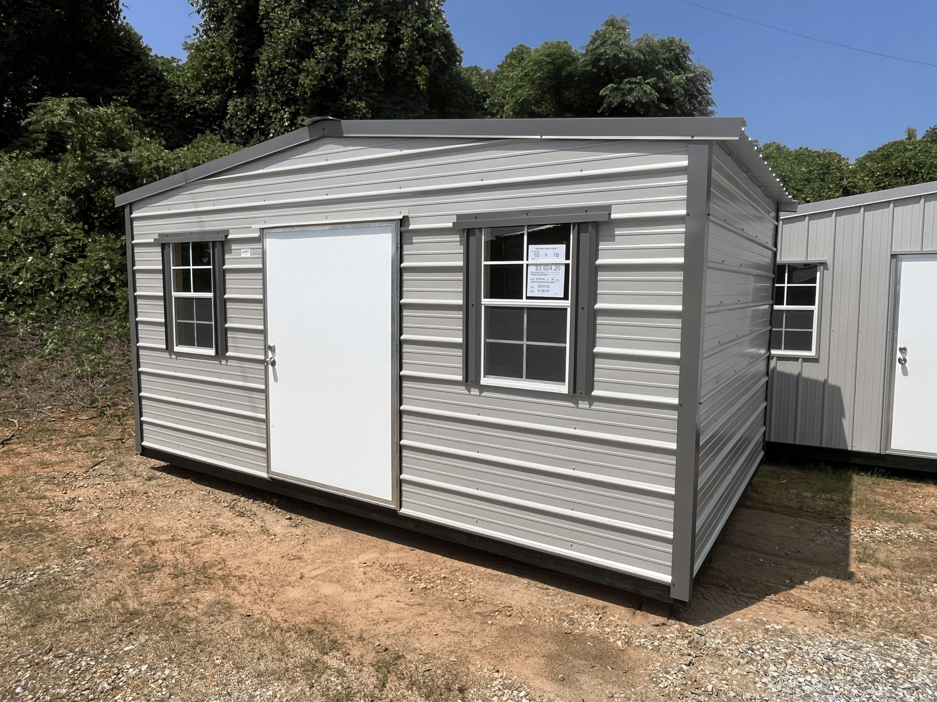 Small gray prefab shed with white door and windows on a dirt lot near trees