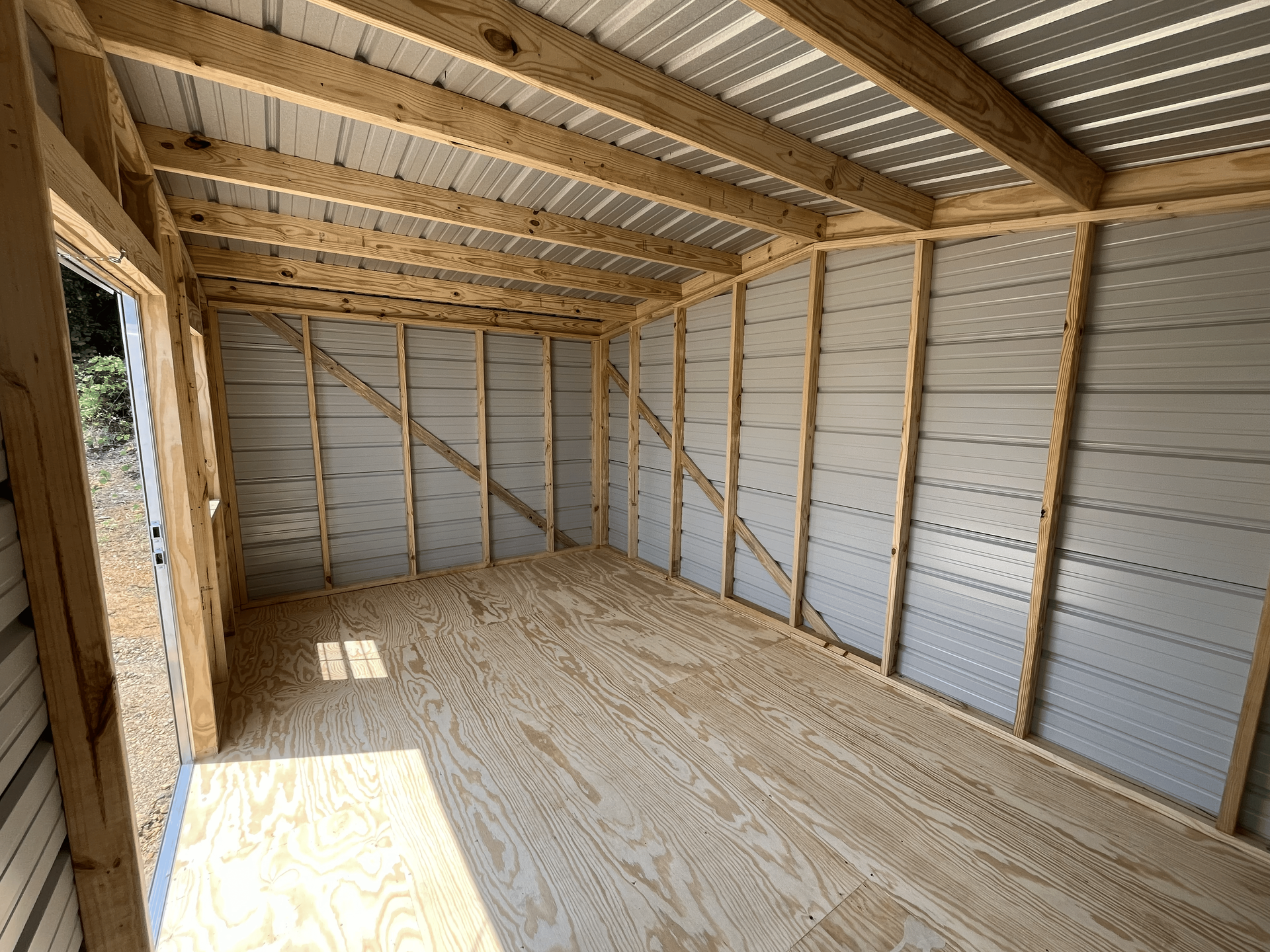 Empty unfinished wooden room with exposed framing and plywood floor, lit by sunlight from a window.