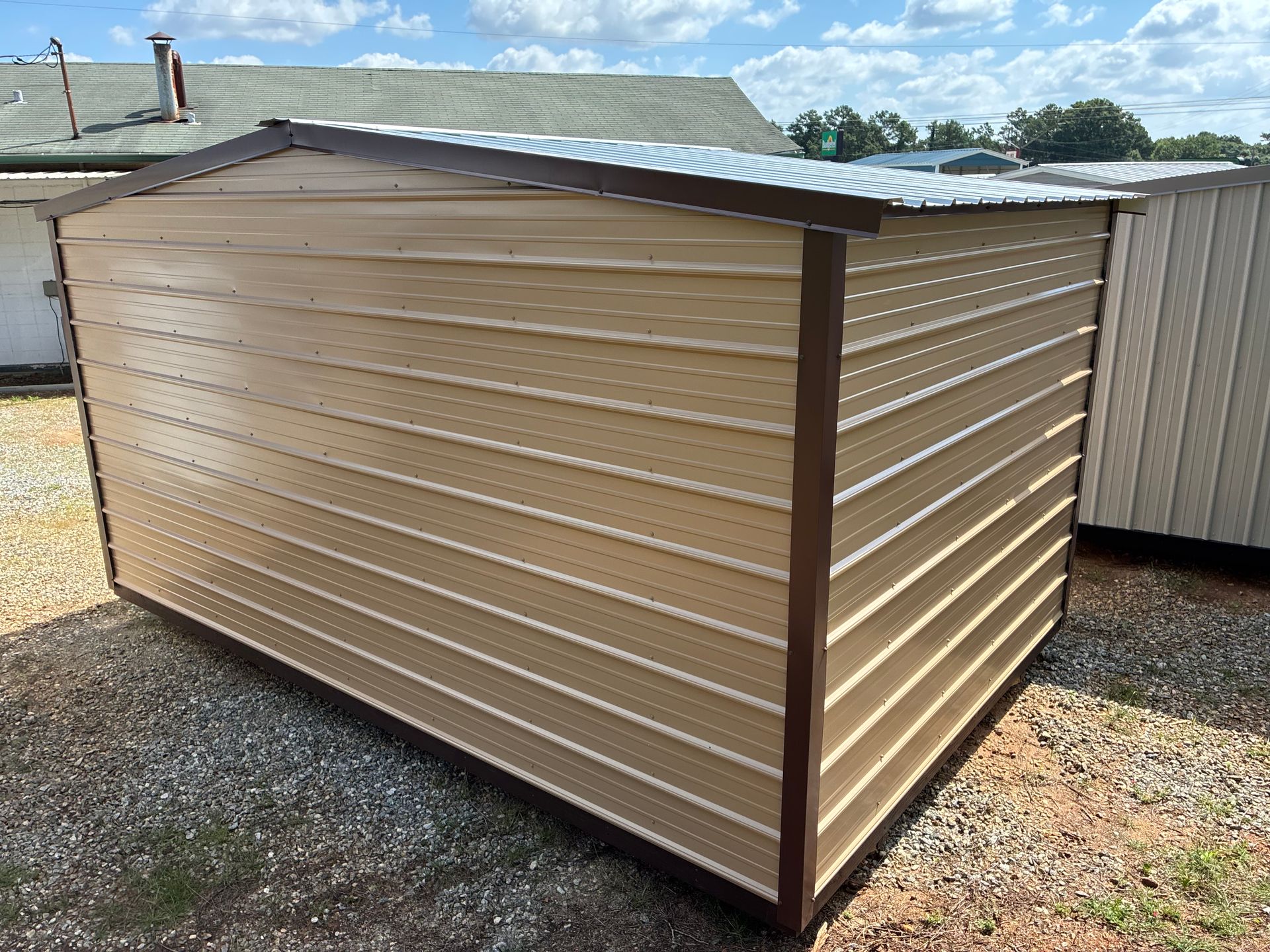 Tan metal shed with corrugated siding and sloped roof on gravel, outdoors beside a larger building