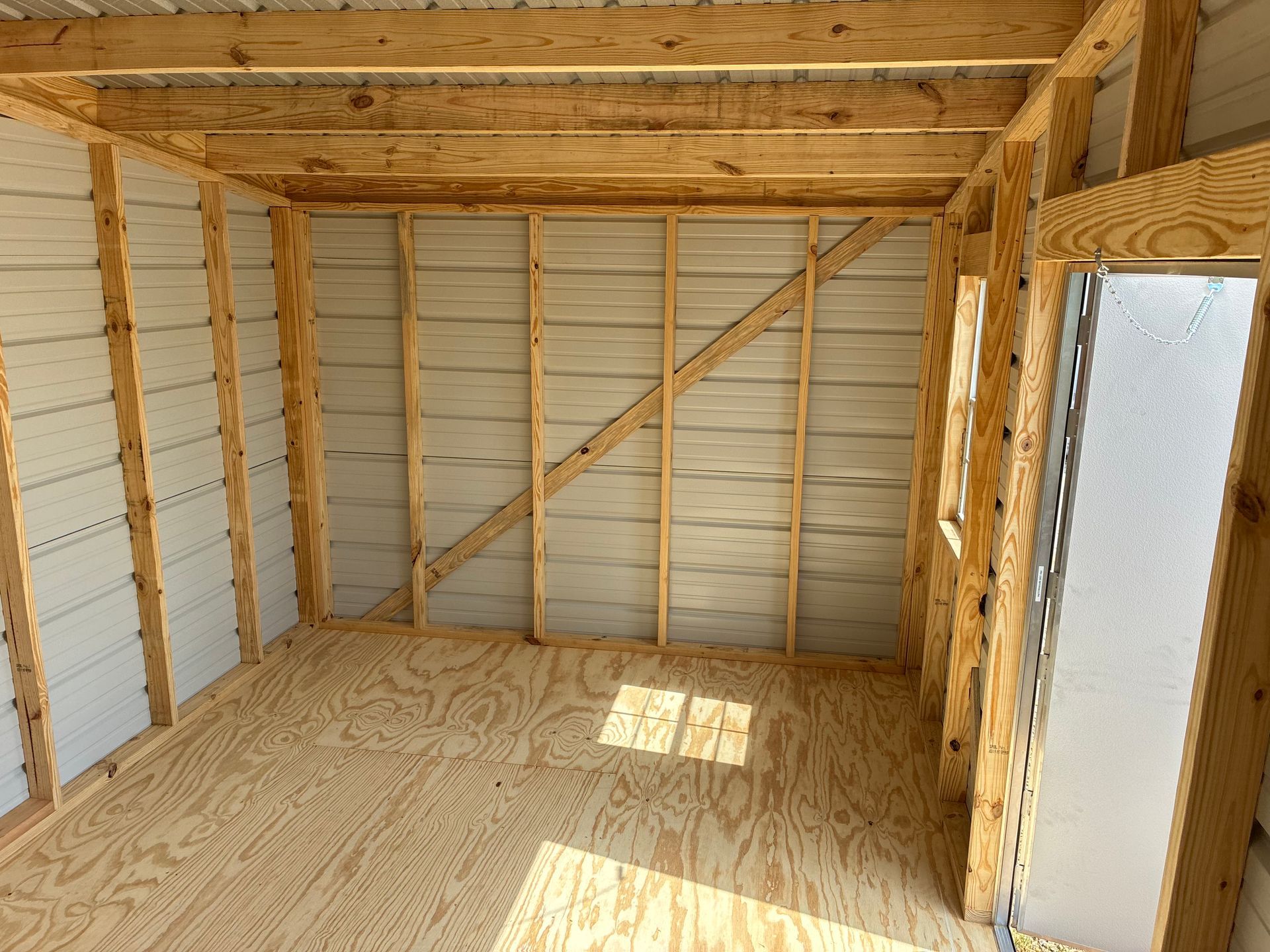Interior of an unfinished wooden shed with exposed framing, plywood floor, and a small windowed door.
