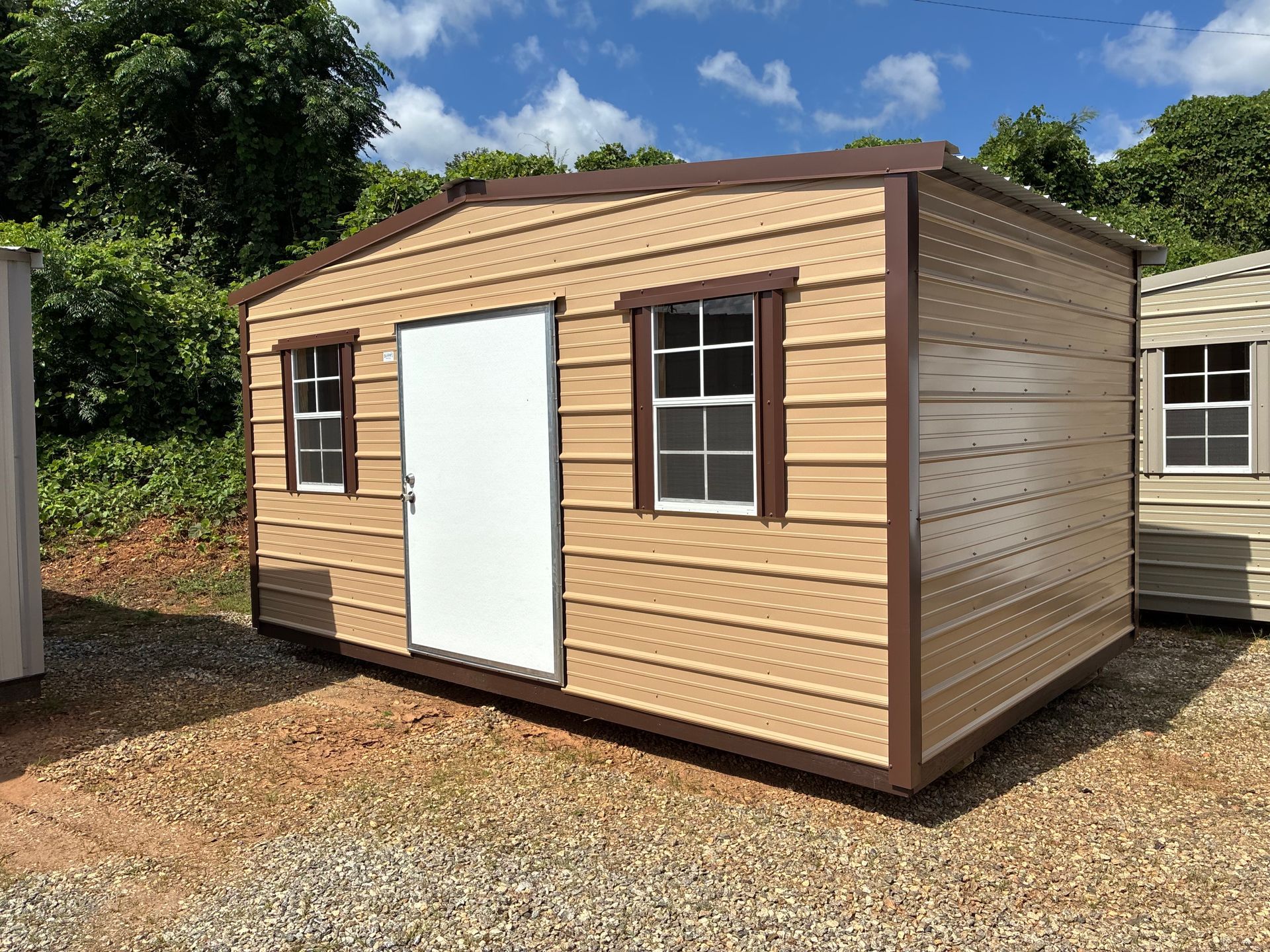 Tan wood storage shed with white door and window, sitting on gravel outdoors under a blue sky.