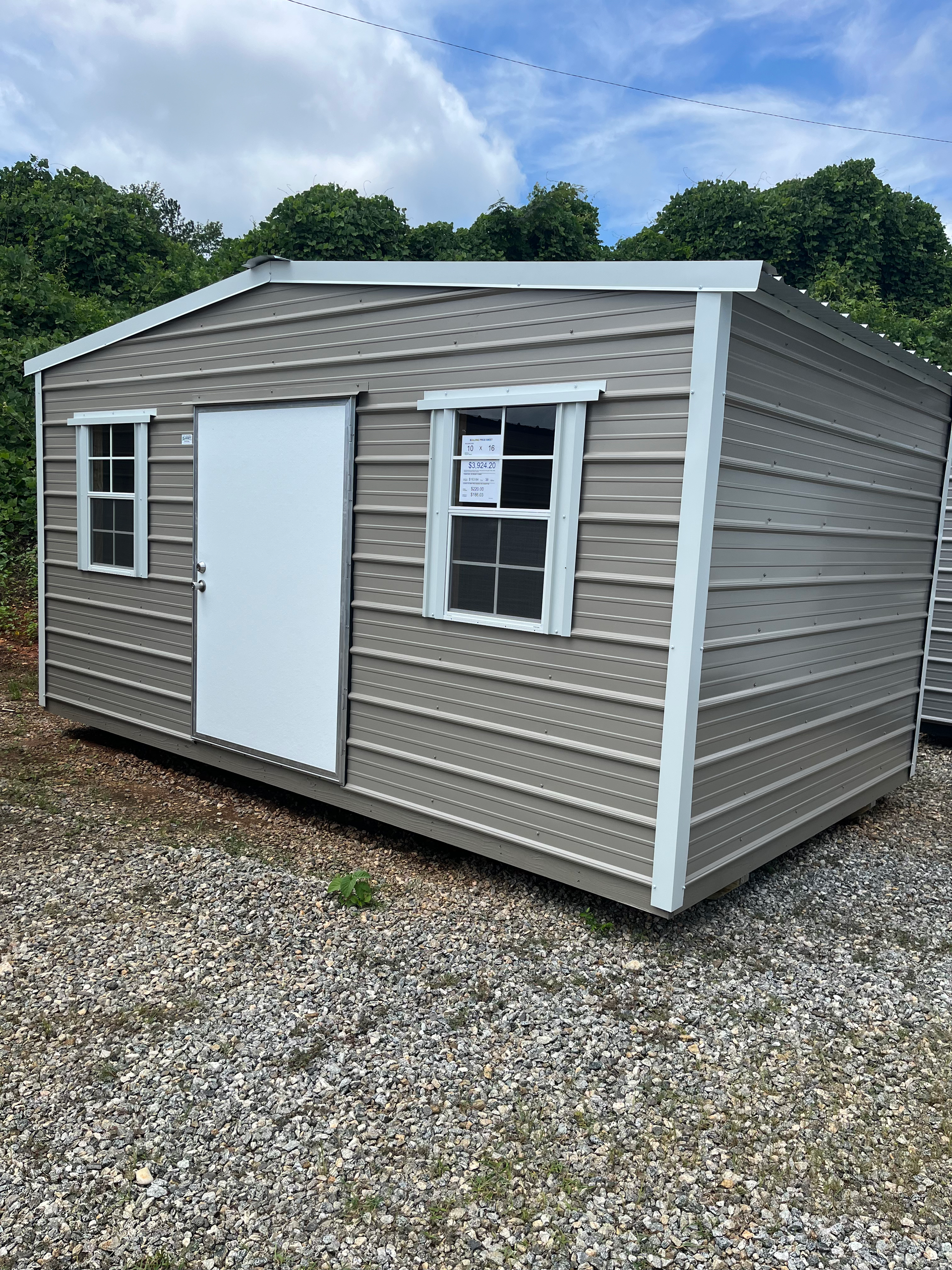 Small beige shed with white trim and door on a gravel lot under a cloudy sky