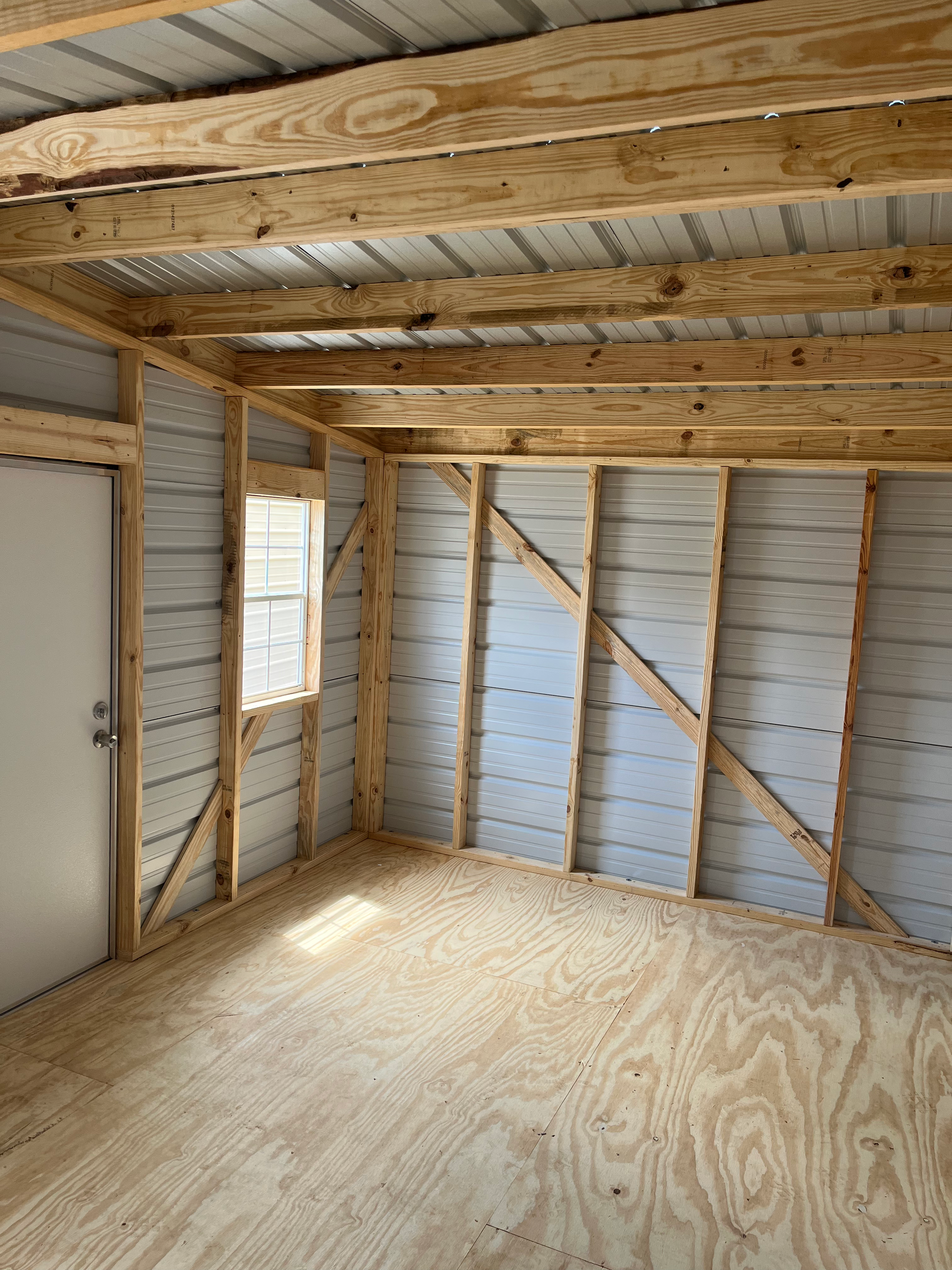 Empty unfinished room with exposed wood framing, plywood floor, and a small window.