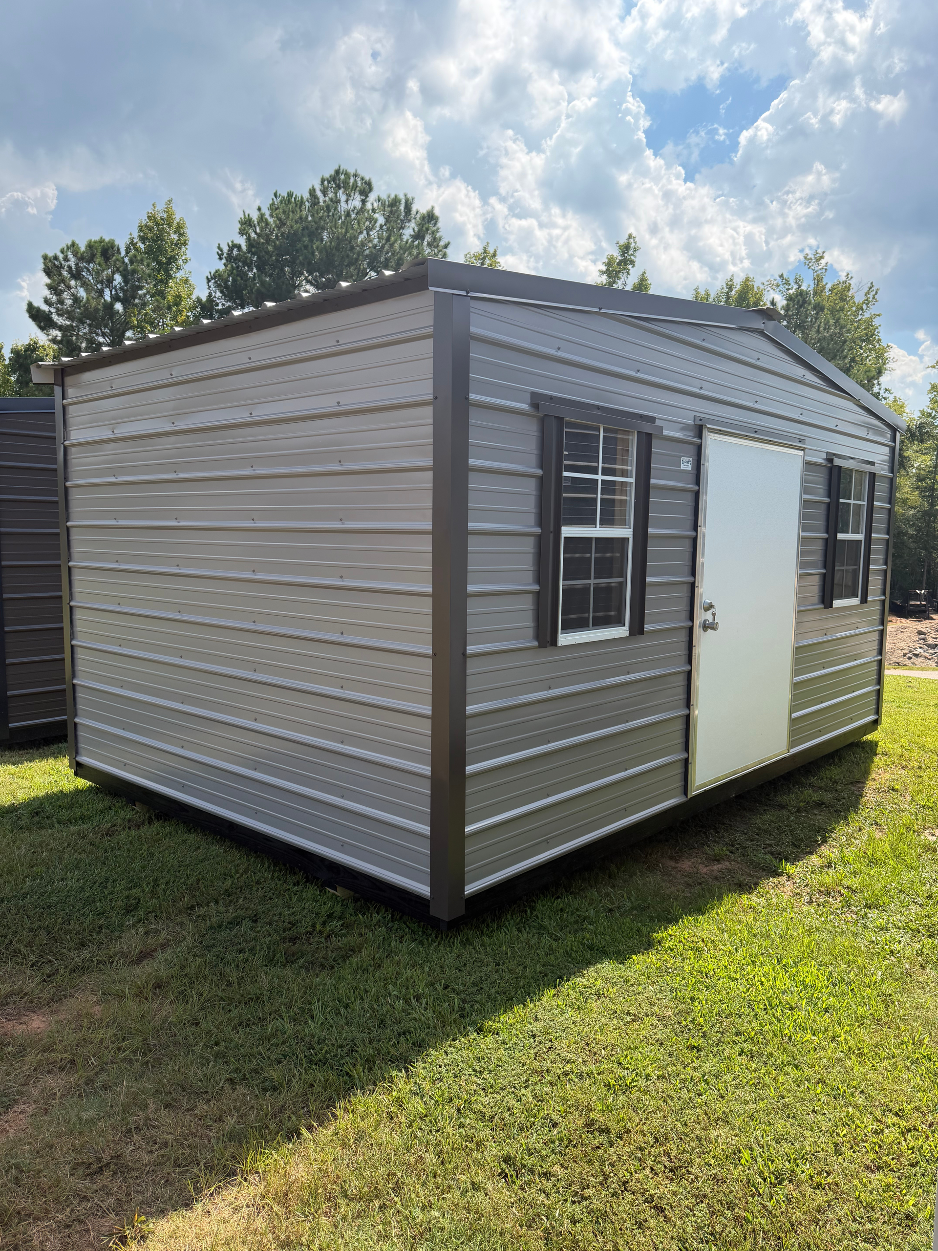 Small gray metal storage shed with a white door and window, sitting on grass under a partly cloudy sky